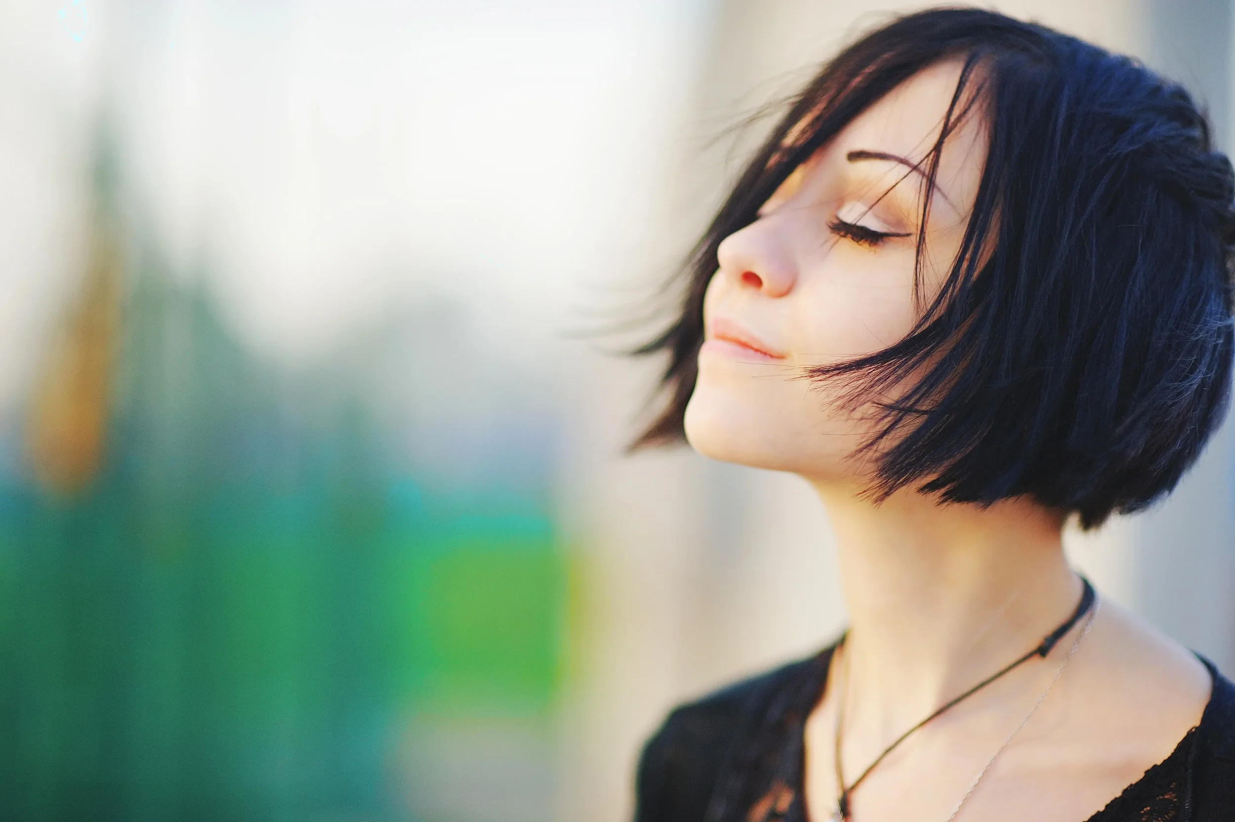 Close-up of a young woman with short, dark hair and closed eyes, smiling softly, outdoors with blurred background.