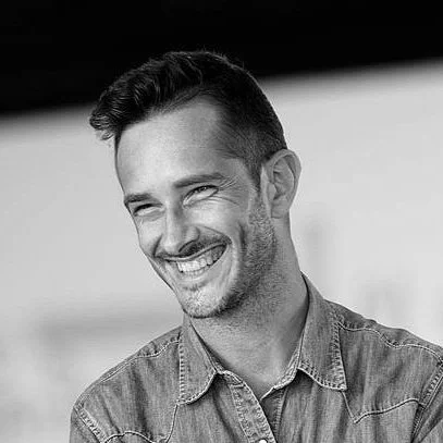 A smiling man with short hair and a beard, wearing a denim shirt, in a black-and-white photo.