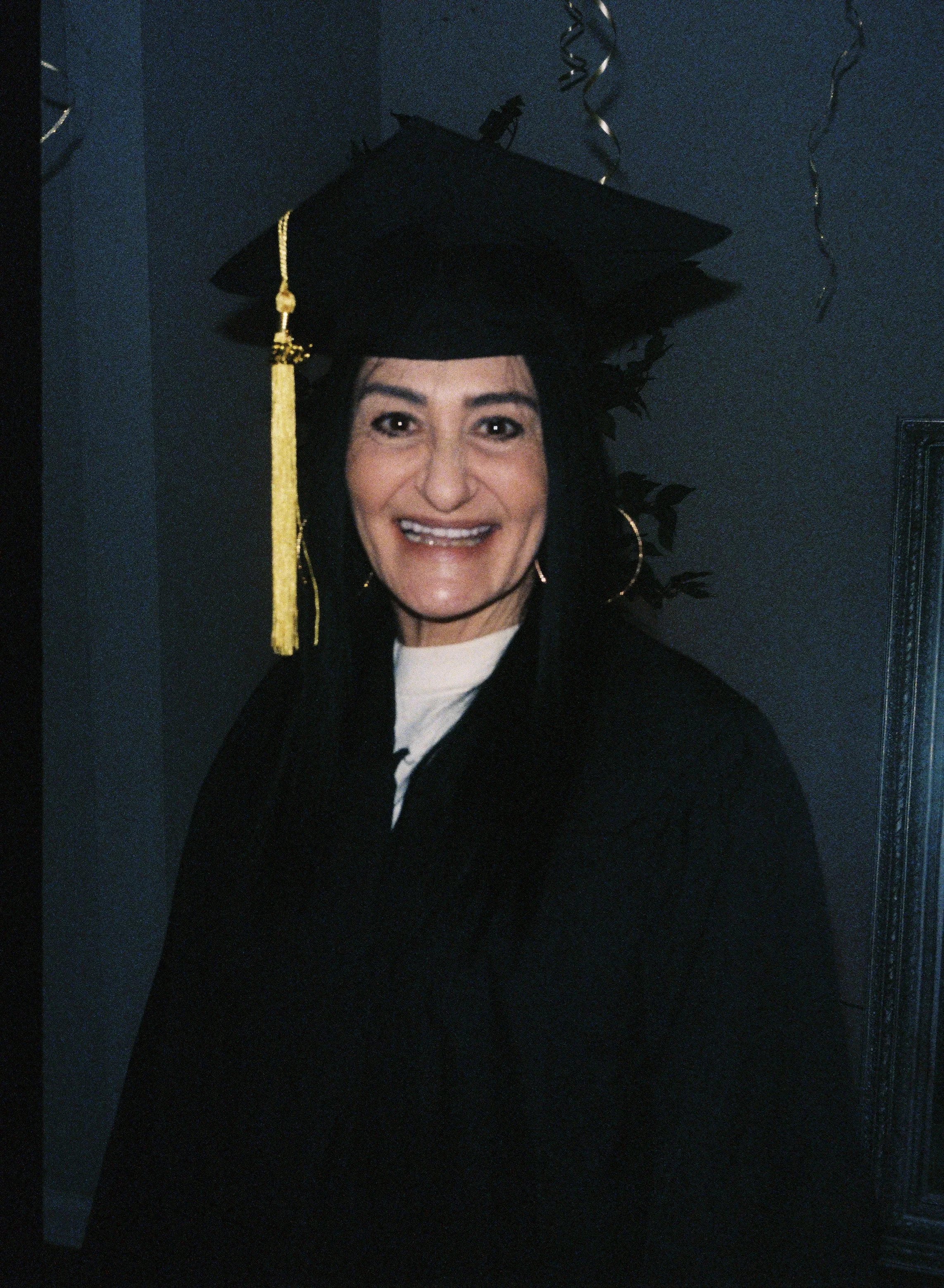 Woman celebrating graduation, wearing a black cap with a yellow tassel and robe, smiling, standing in front of a blue wall with decorative "Happy Graduation" balloon.