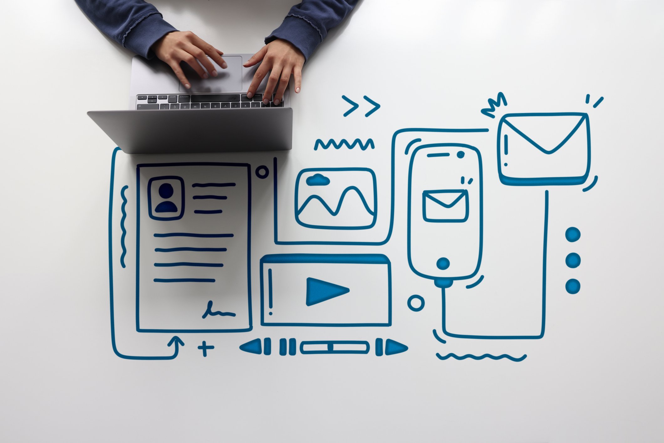 Person working on a laptop at a white table with hand-drawn blue icons depicting social media and communication tools, including profile, images, videos, email, and messaging apps.