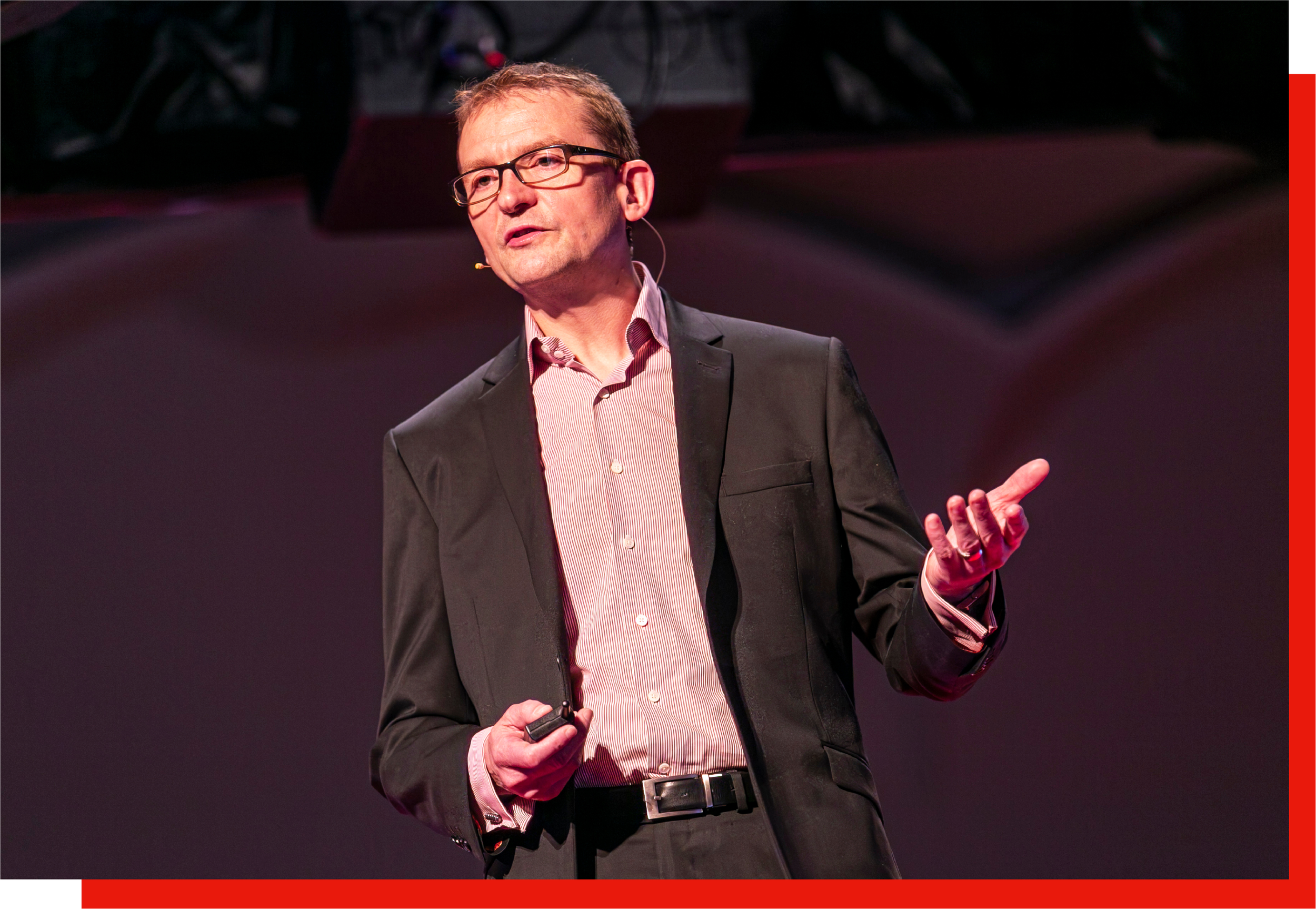 A man in a suit and glasses giving a presentation on stage, holding a device in his right hand.