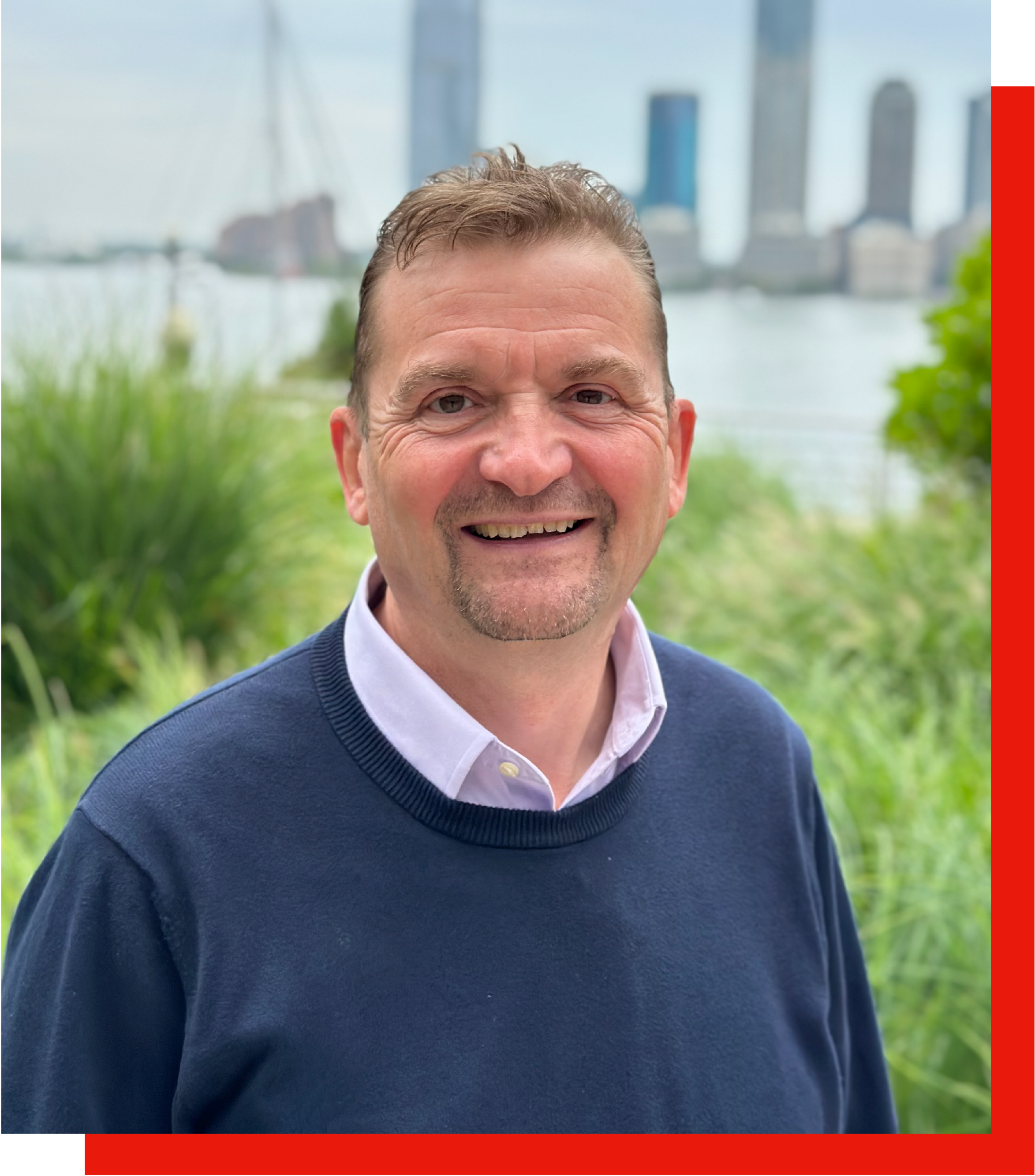 A smiling man with short brown hair, wearing a navy sweater over a light-colored collared shirt, standing outdoors with greenery and a city skyline with tall buildings in the background.
