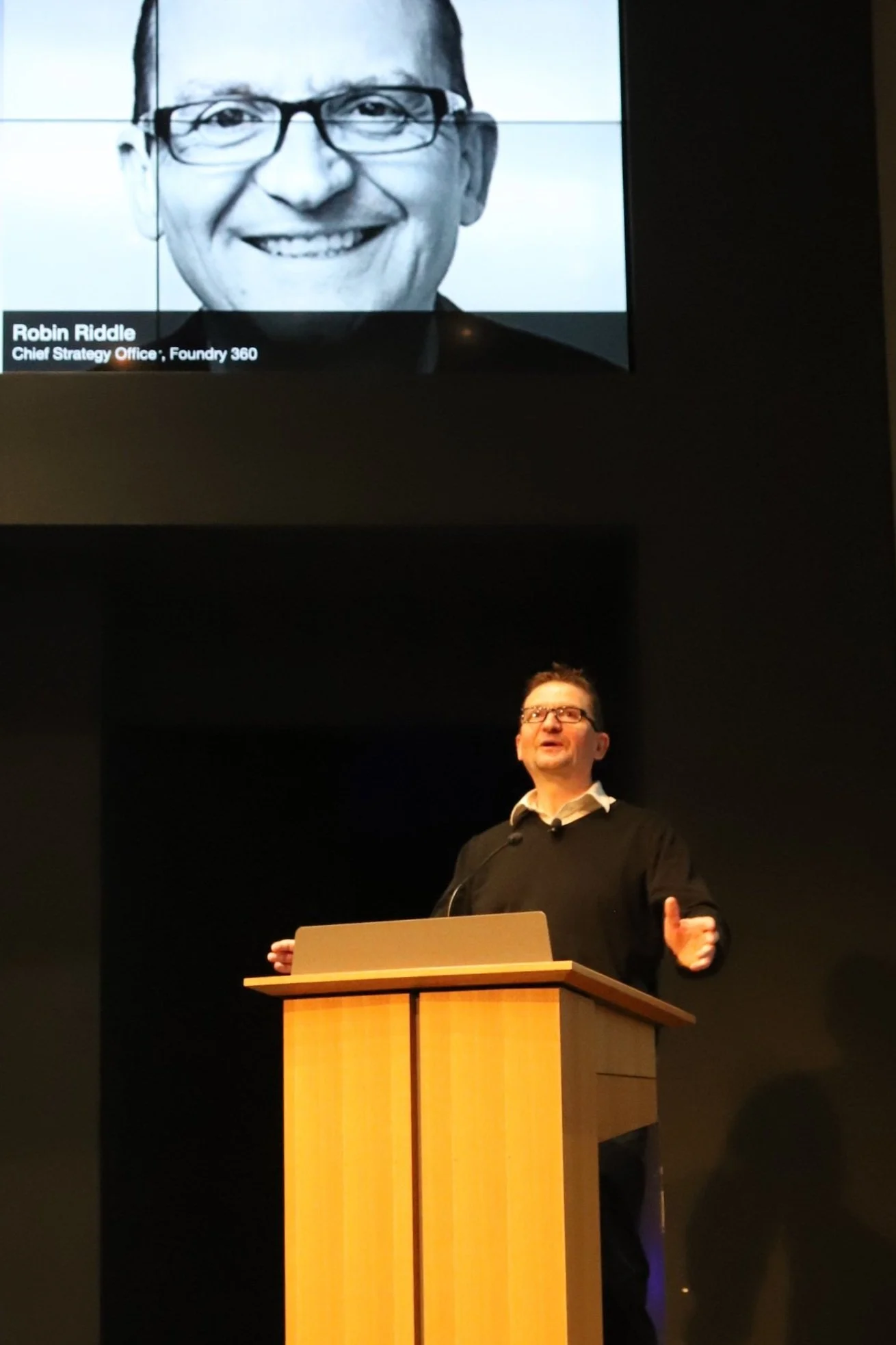 A man wearing glasses and a black sweater standing behind a wooden podium, giving a presentation. A large screen above shows a black and white photo of another man with glasses and a smile, with text that reads "Robin Riddle, Chief Strategy Office, Foundry 360."