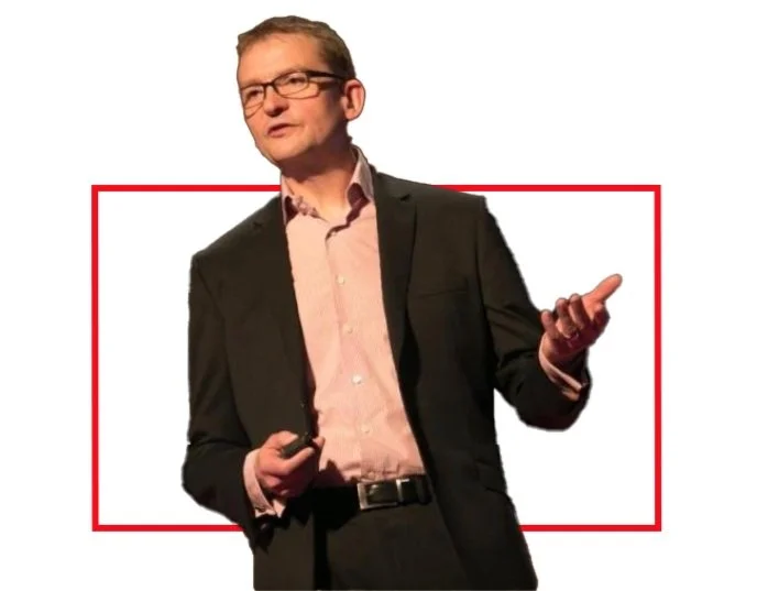 A man in a black suit and glasses giving a presentation on a stage with a white background and a red rectangular border.