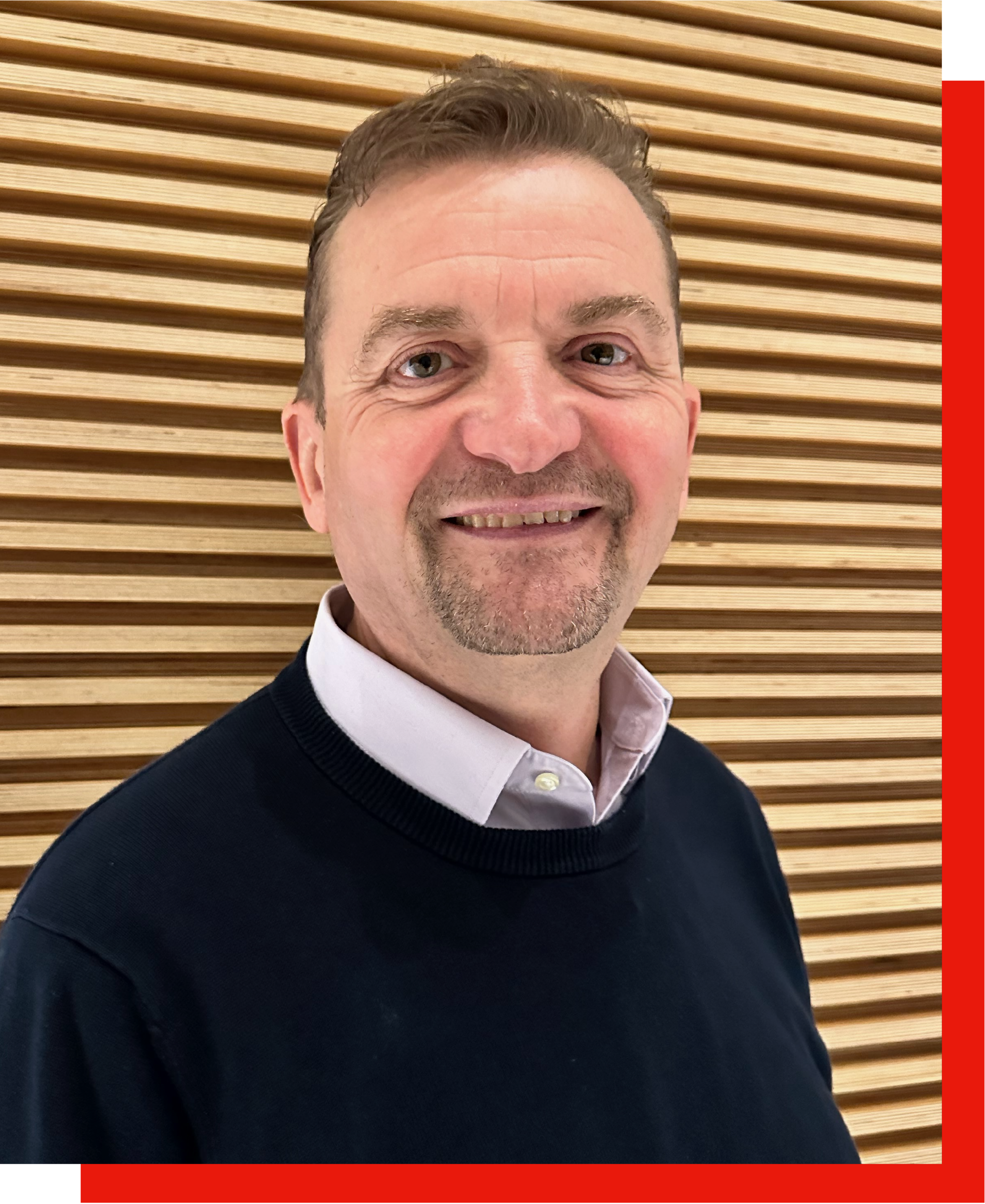 A smiling man with light brown hair and a short beard, wearing a white collared shirt under a dark navy sweater, standing in front of a wood panel wall.