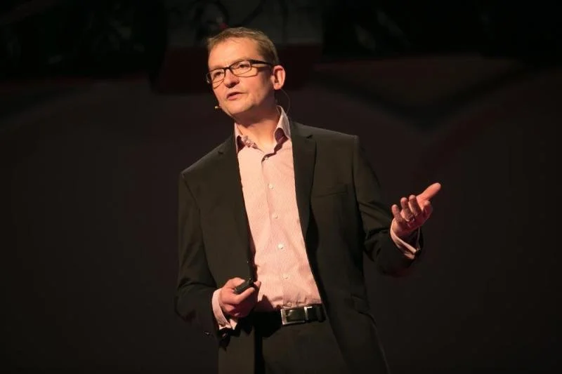 Man in a suit giving a presentation on stage with a microphone in his hand, dark background.
