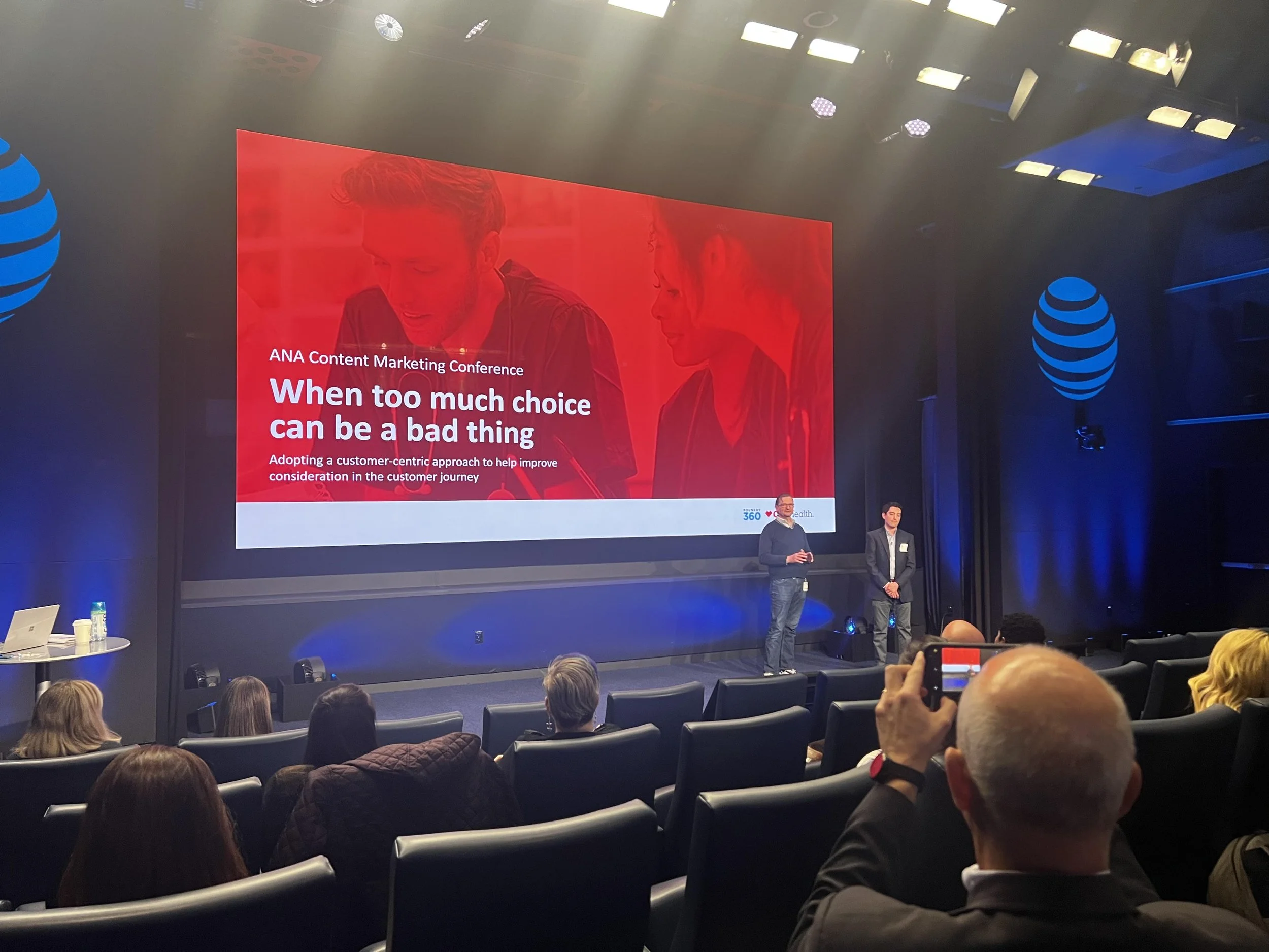 A conference room with a large screen displaying a presentation titled 'When too much choice can be a bad thing' at the ANA Content Marketing Conference. There are two speakers on stage, one in a dark sweater and jeans, and the other in a dark suit, addressing an audience seated in black chairs. The room has blue lighting with the AT&T logo on the walls.