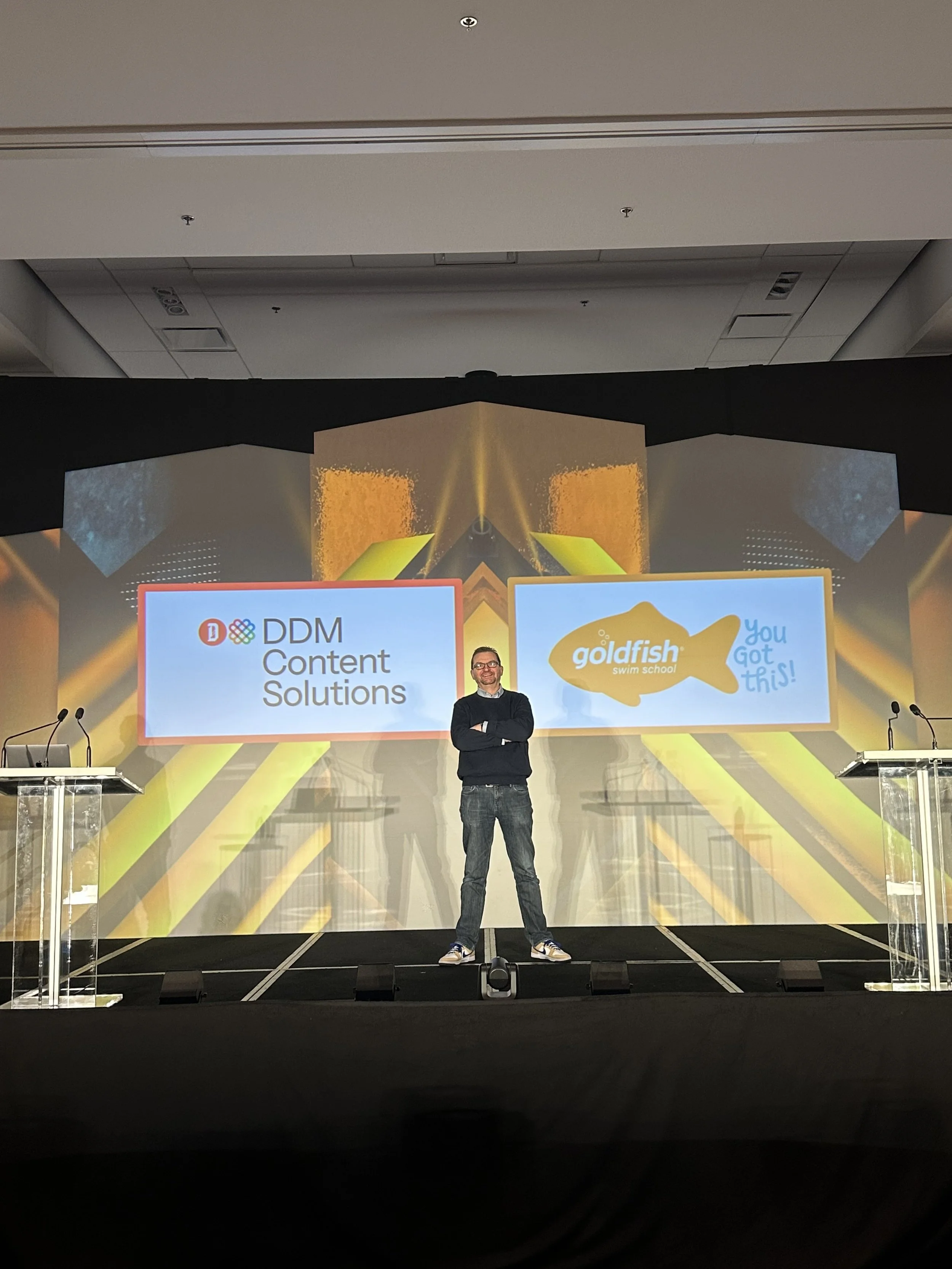 A man standing on a stage in front of large screens displaying two logos, one for DDM Content Solutions and one for Goldfish Swim School, with the man smiling and arms crossed.