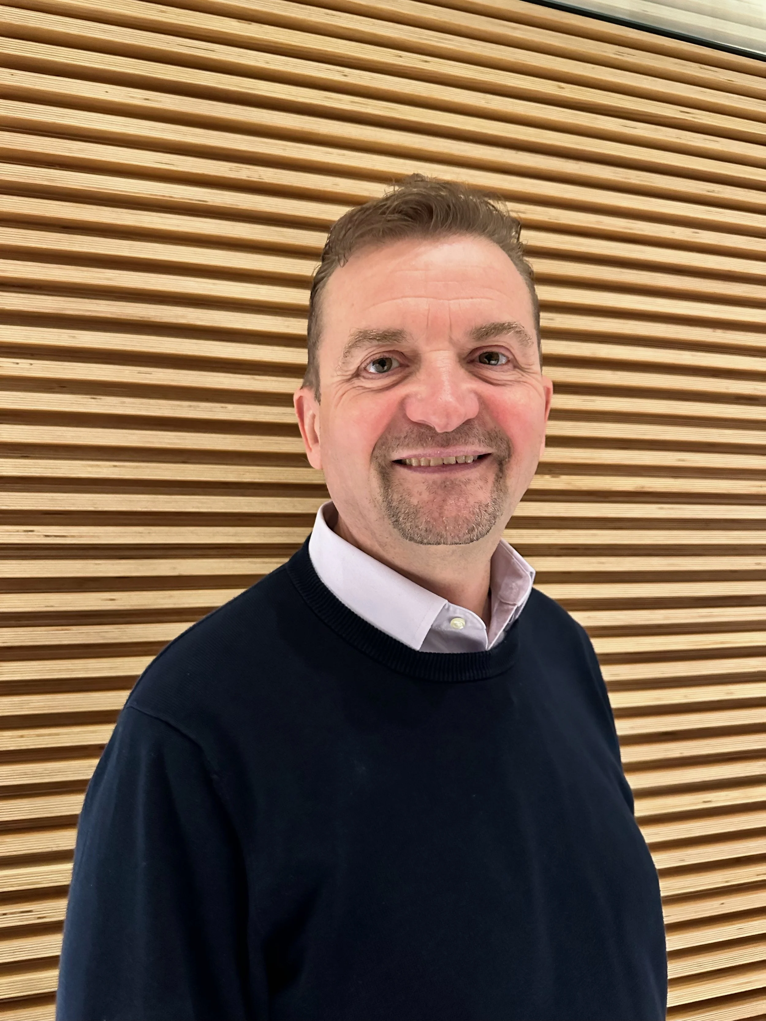 A middle-aged man with light skin, short brown hair, and a goatee, wearing a white collared shirt and a black sweater, smiling in front of a background with horizontal wooden slats.