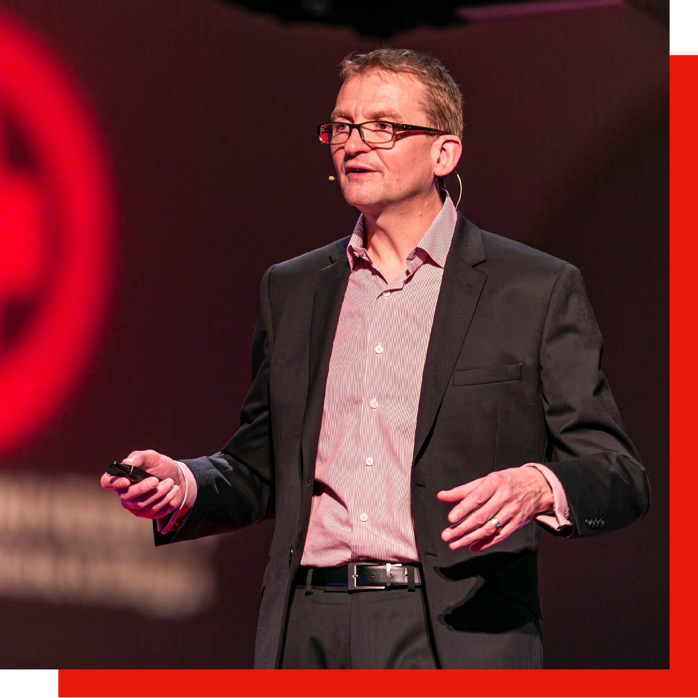 A man wearing glasses and a black suit giving a presentation on stage, holding a remote control in his right hand, with a dark background.