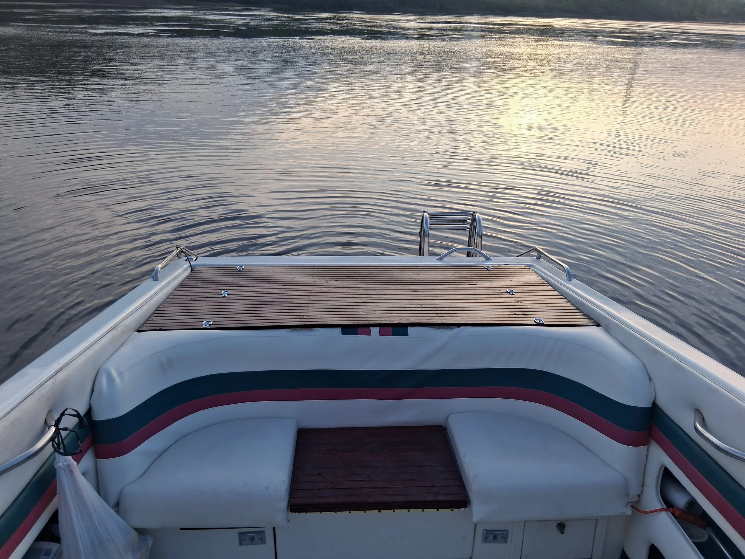 The image features a view from the front of a boat on calm water during sunset or sunrise, with reflections of light on the water surface, and a metal ladder at the back of the boat.