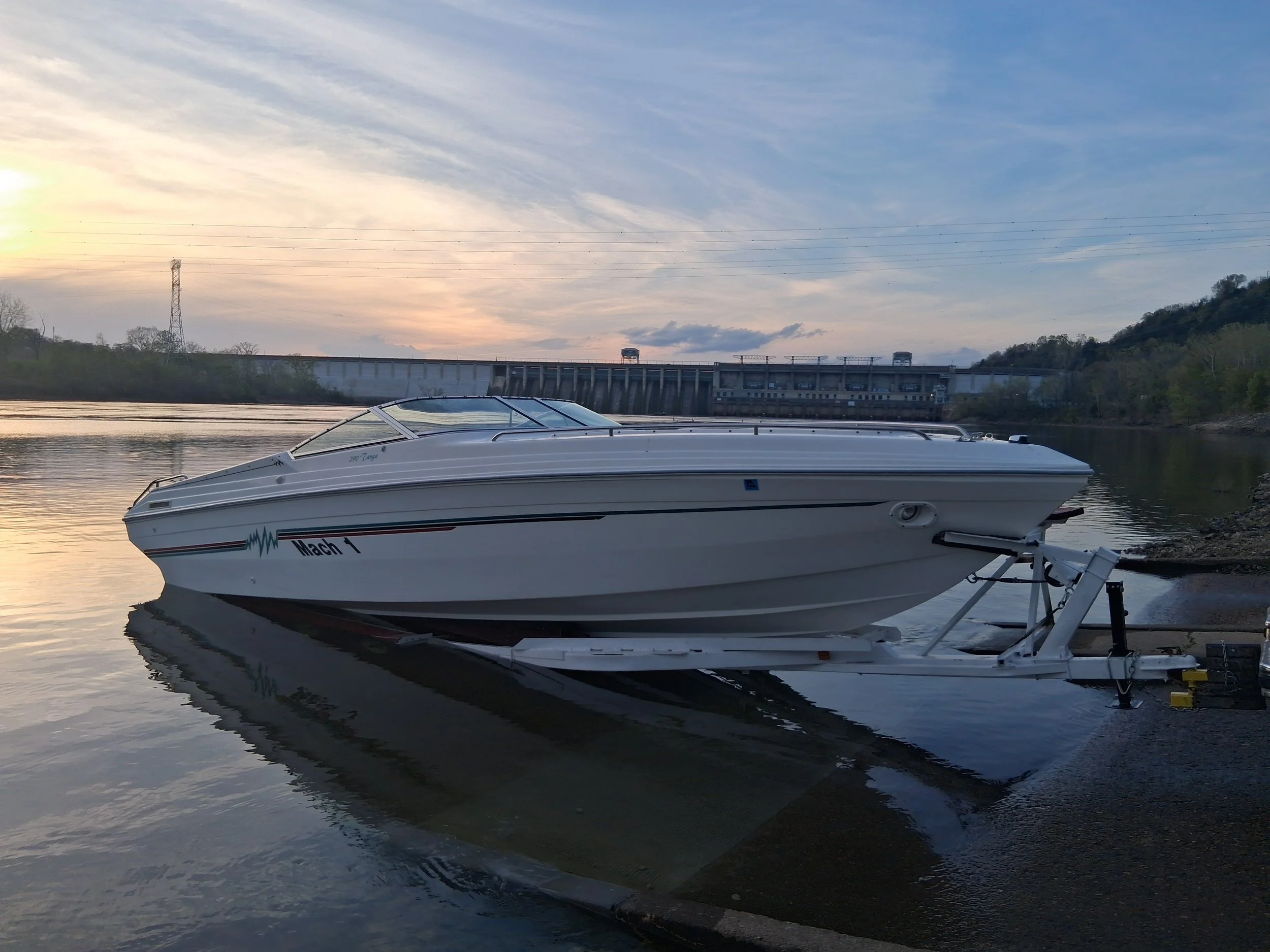 A white speedboat named Mach 1 is docked on a calm river with a dam and rolling hills in the background during sunset or sunrise.