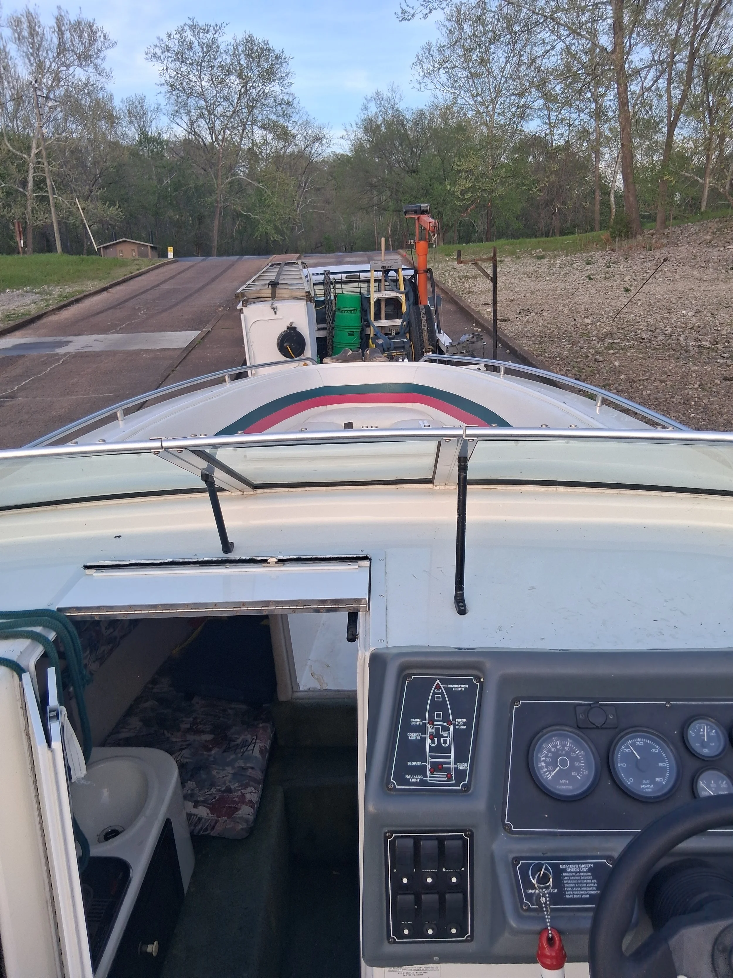 View from the driver's seat of a boat showing the dashboard, steering wheel, and controls, with a trailer carrying a boat attached to a vehicle on a road surrounded by trees and grass.