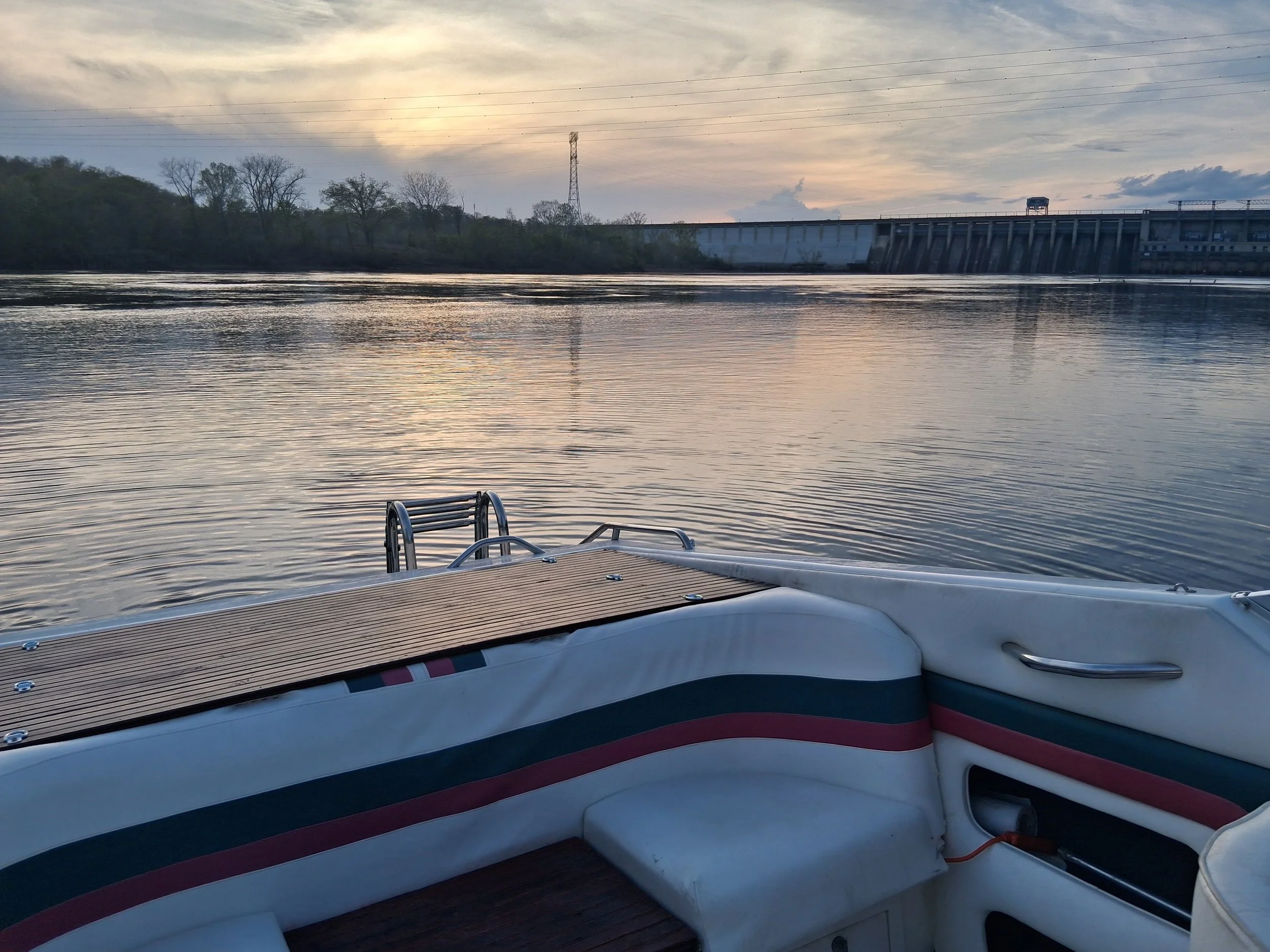 View from a boat on a river at sunset, with a dock or boat deck in the foreground and trees, a dam, and a cloudy sky in the background.