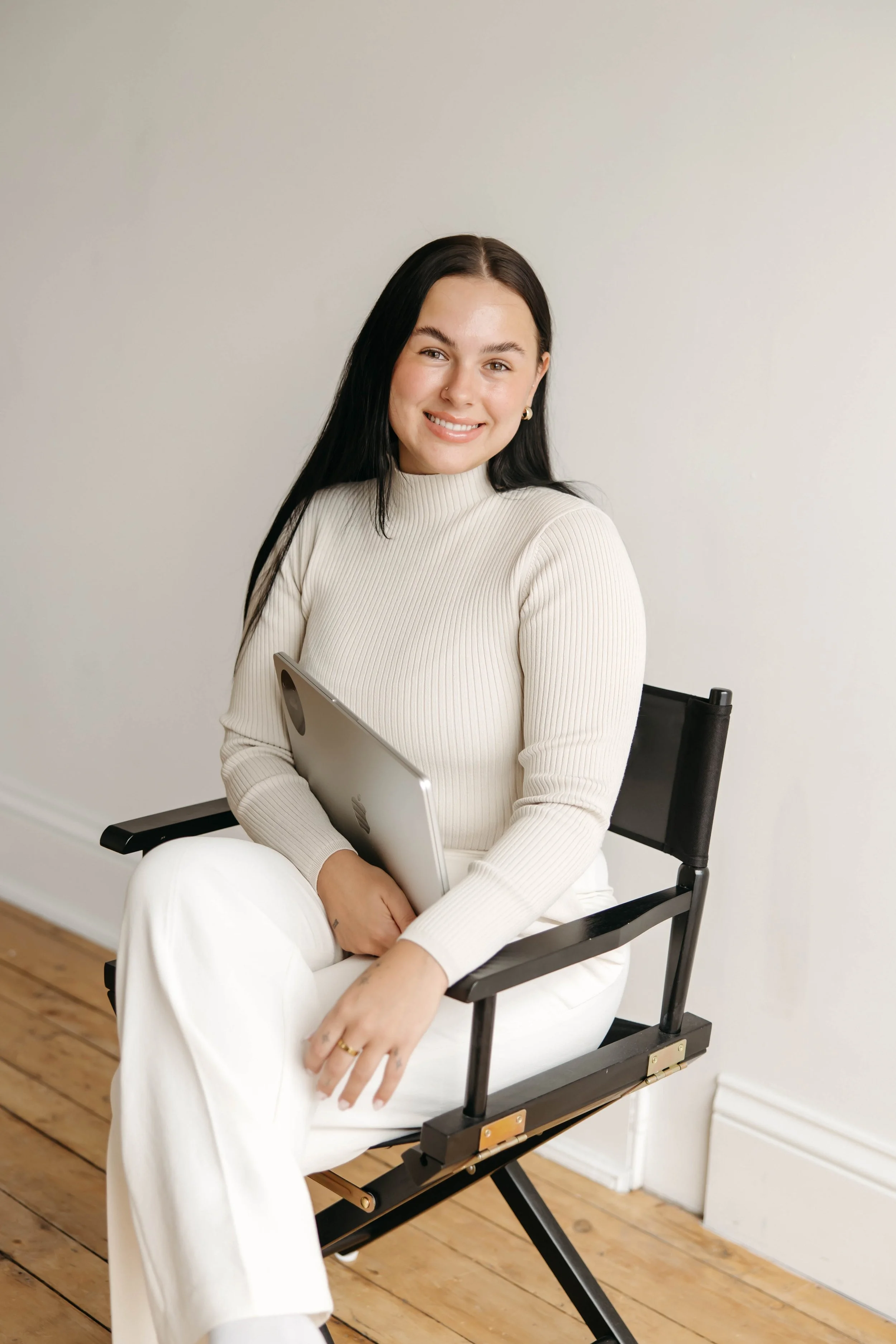 A woman sitting in a black director's chair on a wooden floor, holding a silver laptop, smiling at the camera, wearing a cream sweater and white pants.