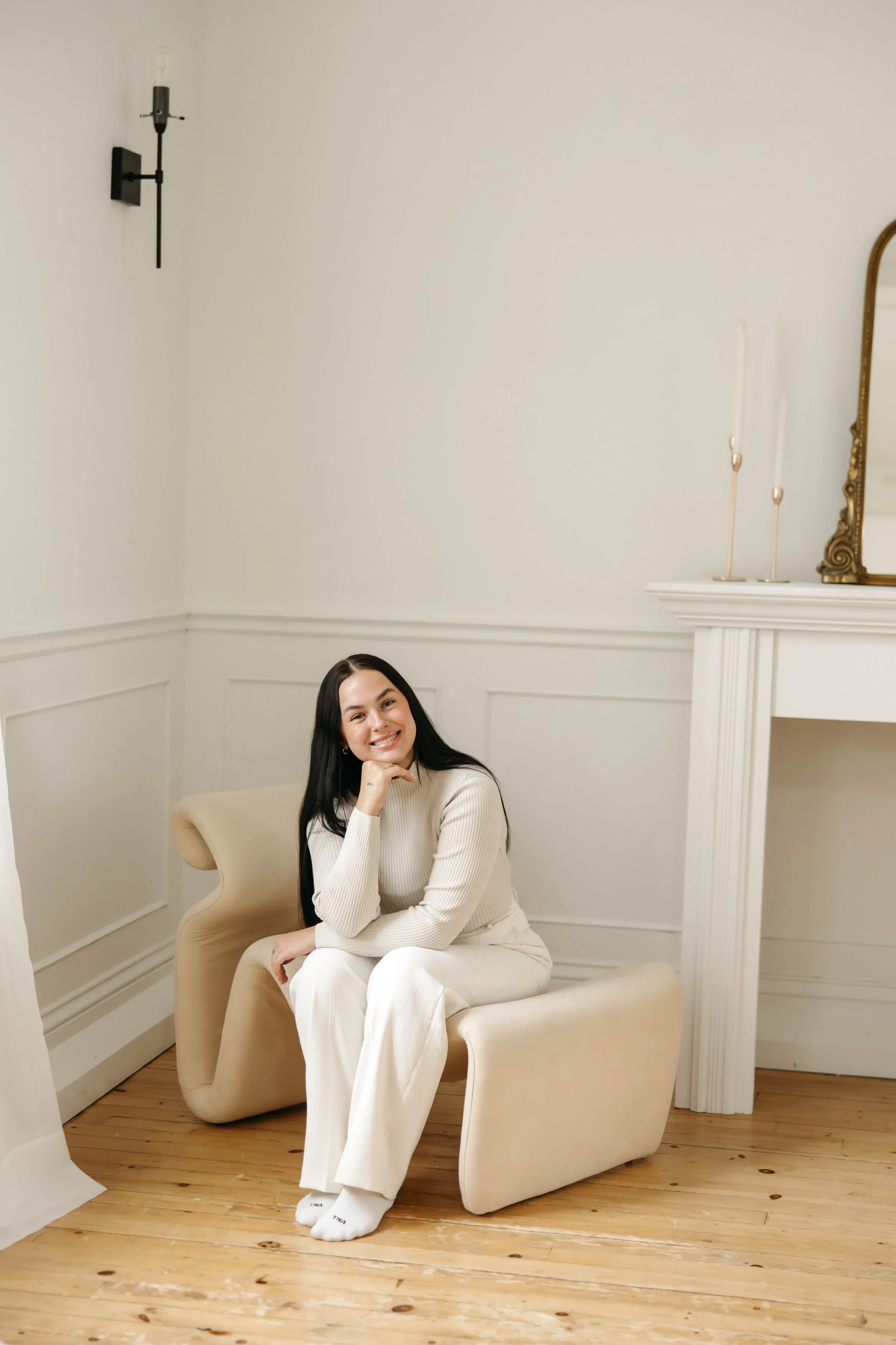 A woman with long dark hair sitting on a modern beige armchair in a white room, smiling, wearing a light beige sweater and white pants.