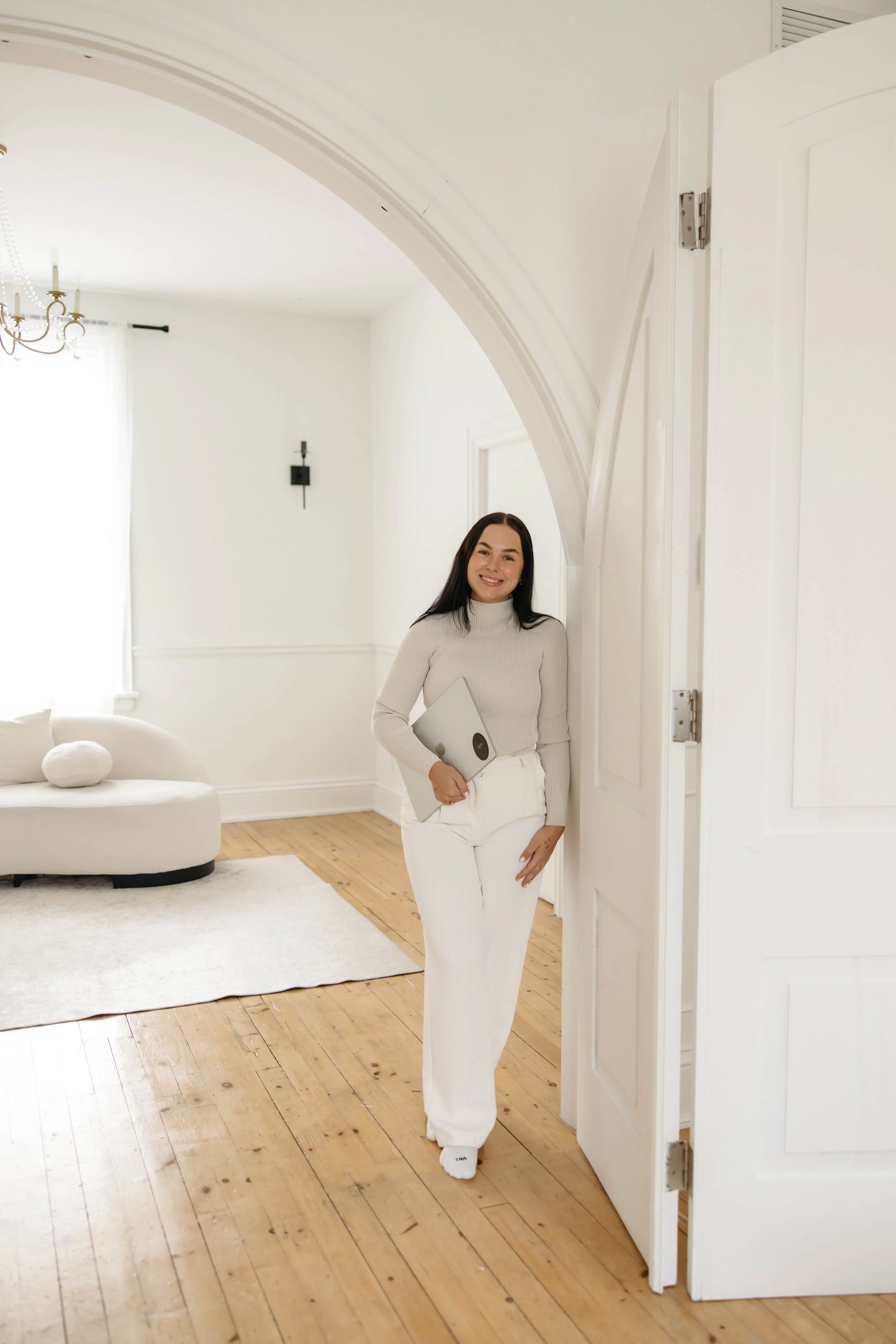A woman with long dark hair smiling and holding a laptop, standing in a bright, white-walled room with wooden floors, a white sofa with a decorative pillow, and a chandelier.