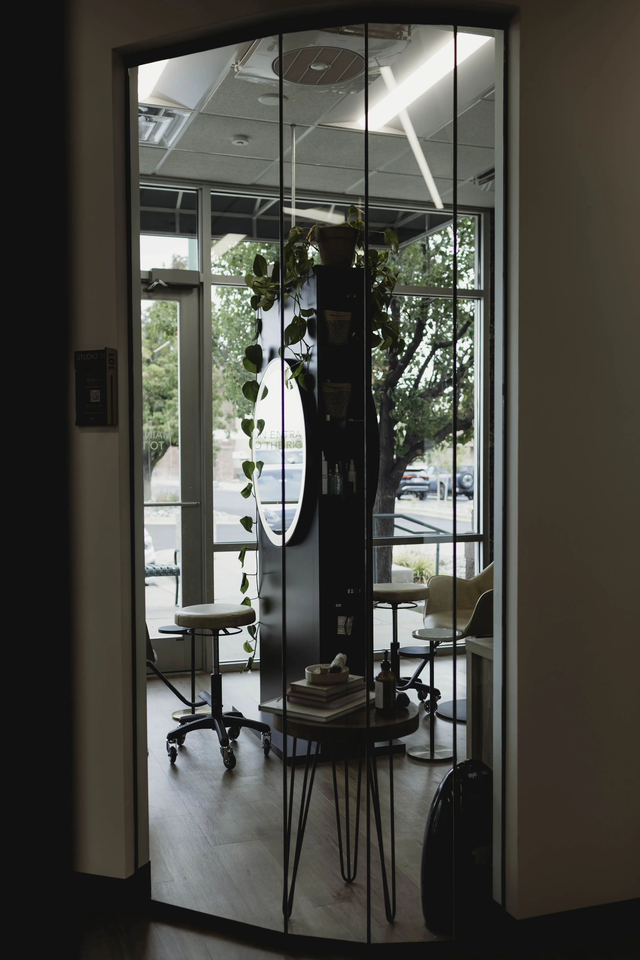 Interior view of a beauty salon or studio, seen through a glass door with black frame, showing salon chairs, shelves with products, and a large window with trees outside.