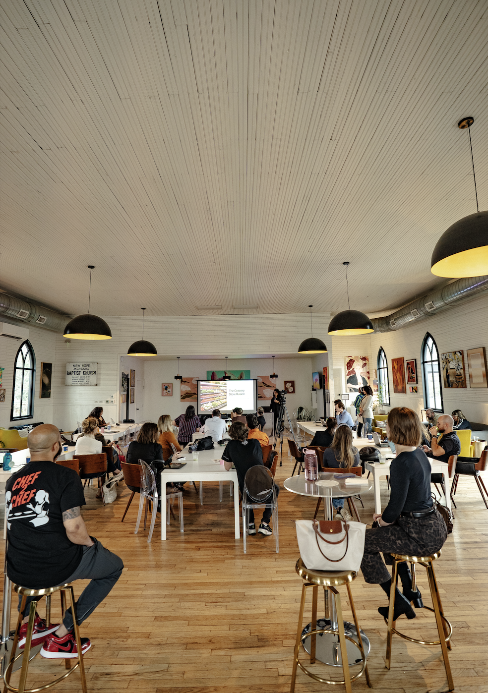 A group of people attending a presentation or workshop in a bright, spacious room with high ceilings, wooden floors, and large arched windows. The room is decorated with various artworks on the walls and has black hanging pendant lights. Some attendees are seated at white tables, while others are standing or seated on bar stools, and a projector screen displays a slide titled 'The Grocery Store Business'.