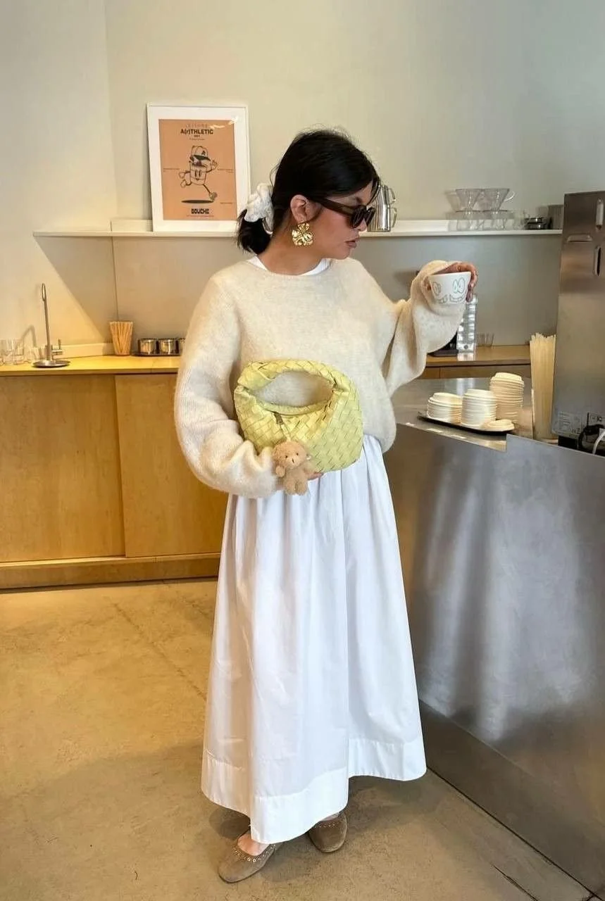Woman wearing large sunglasses and earrings, holding a patterned yellow handbag with a plush toy attached, standing in a cafe with a white skirt and cream sweater, drinking from a cup.