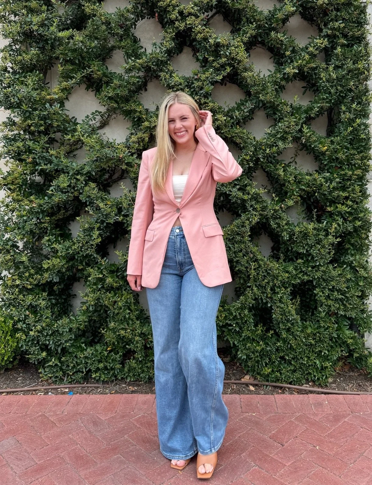 A young woman with long blonde hair is smiling and posing in front of a lush green shrub with a star-shaped pattern. She is wearing a pink blazer, white top, blue jeans, and beige open-toe heels.