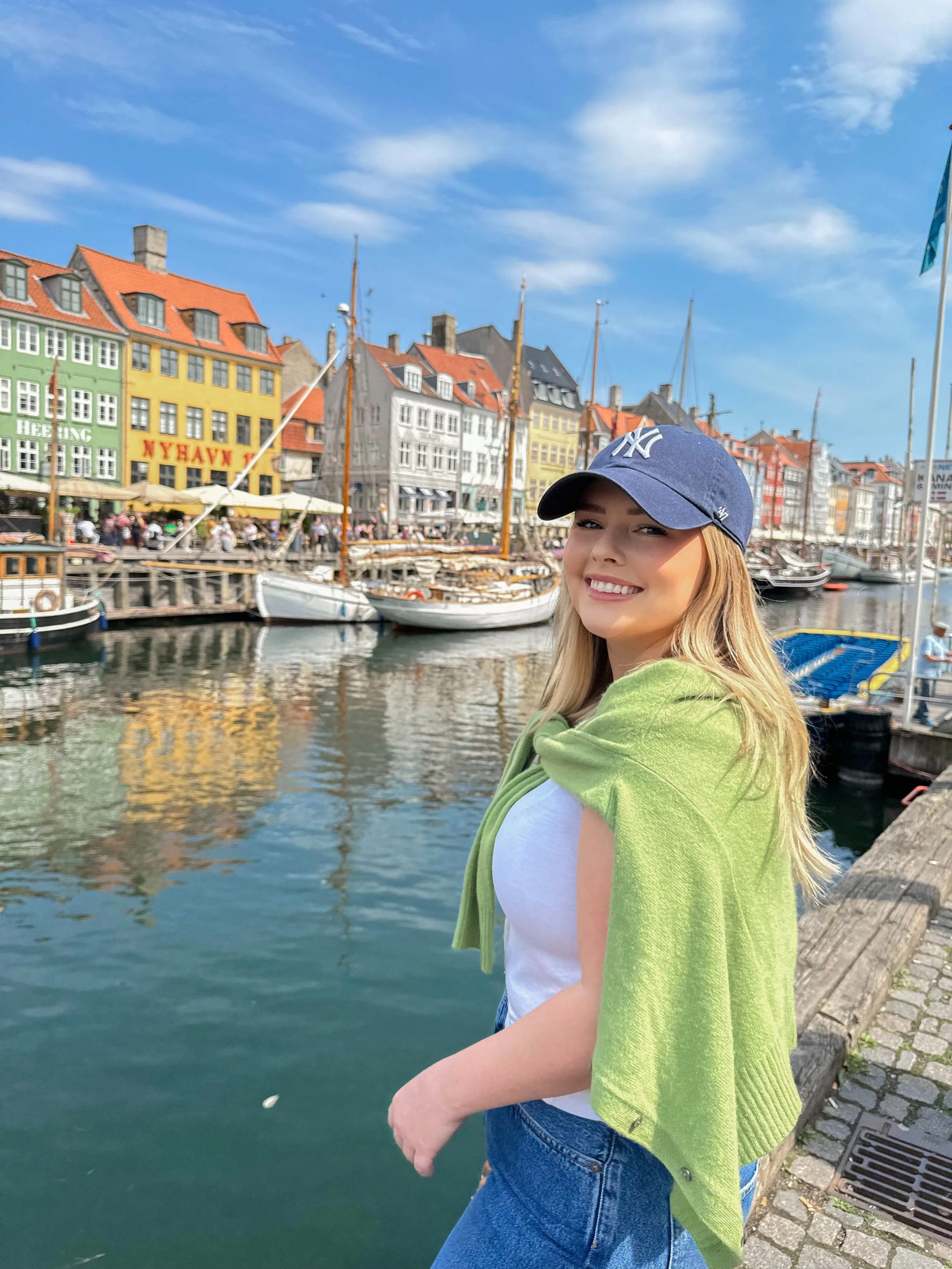 A smiling young woman with blonde hair wearing a navy baseball cap, a white t-shirt, and a green jacket draped over her shoulders, standing by a harbor with boats and colorful buildings in the background.