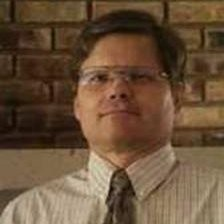 An image of the author, a man wearing glasses, a dress shirt, and a tie standing against a brick wall.