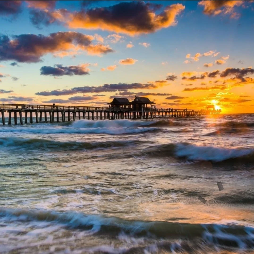 A pier extends into the ocean at sunset, with waves crashing against the shore and clouds in the sky illuminated by the setting sun.