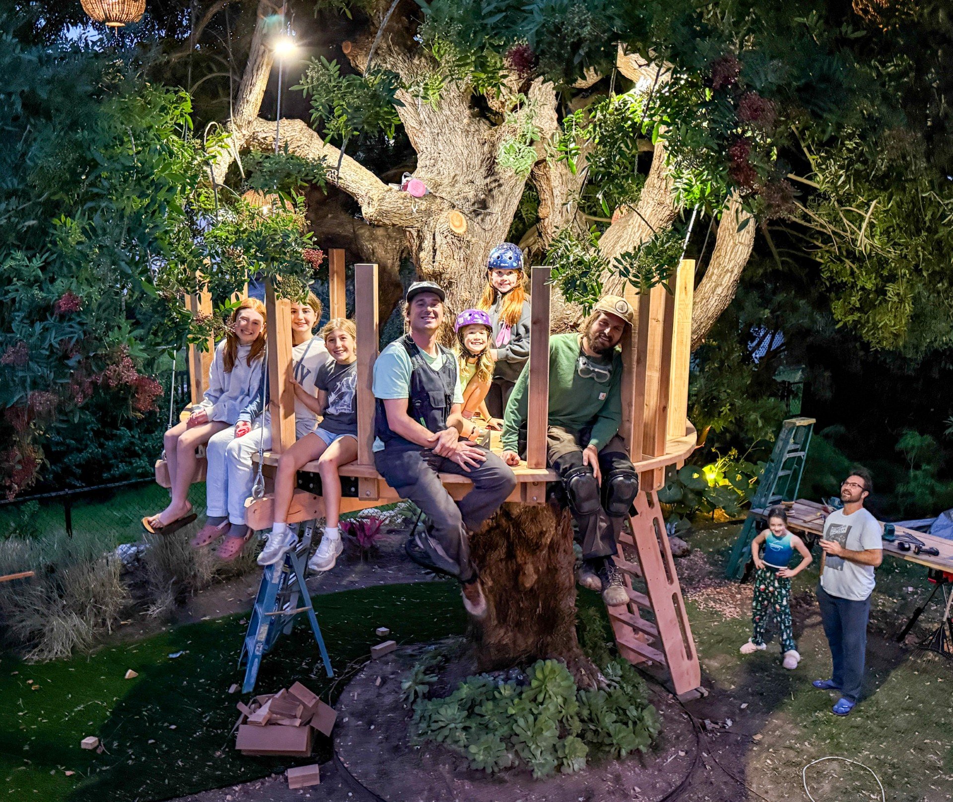 Group of people on a treehouse in a backyard with string lights and shrubbery, some sitting and some standing, with two adults and children wearing helmets and casual clothes.