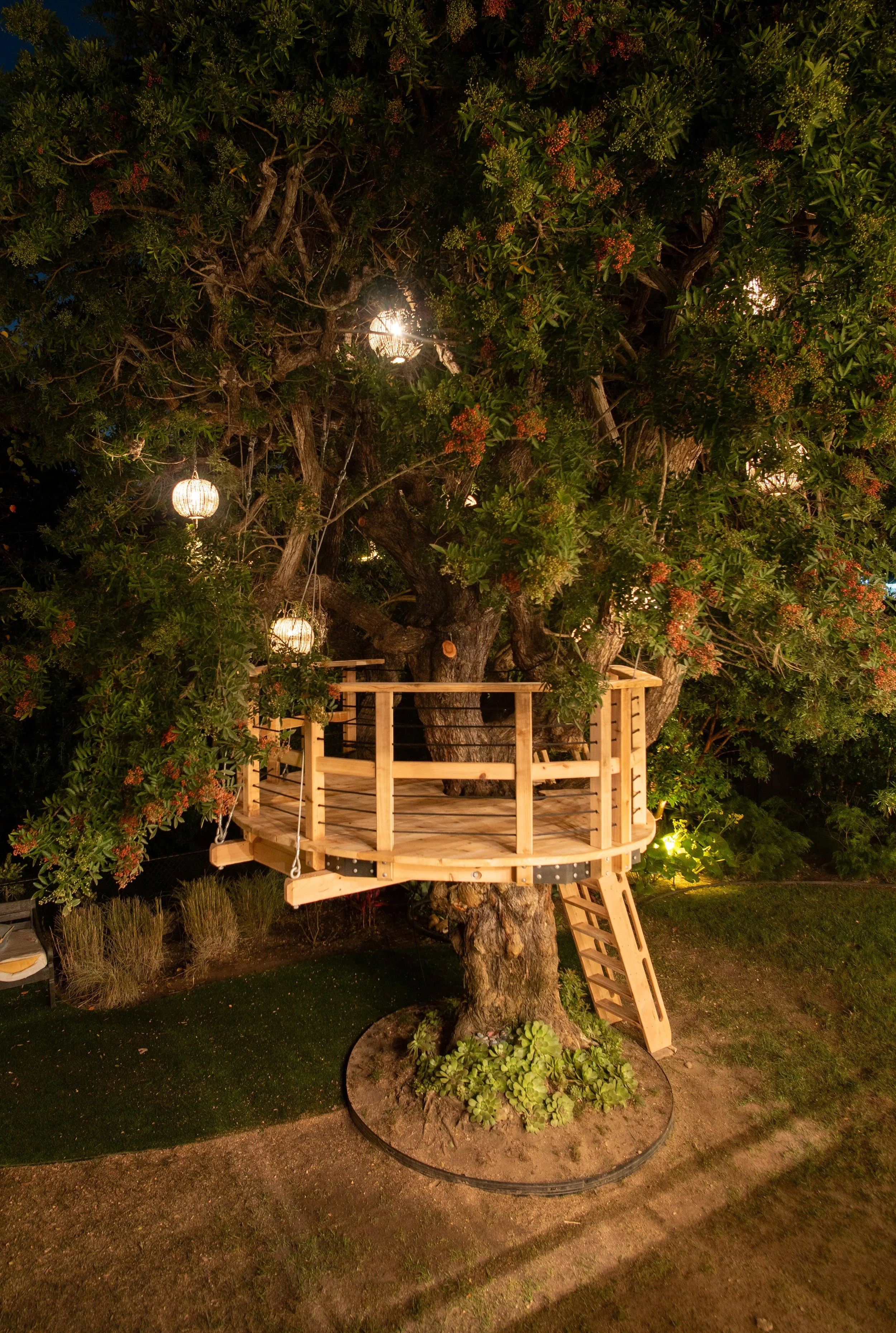 A large tree with an elevated wooden treehouse or lookout platform and hanging lanterns at night.