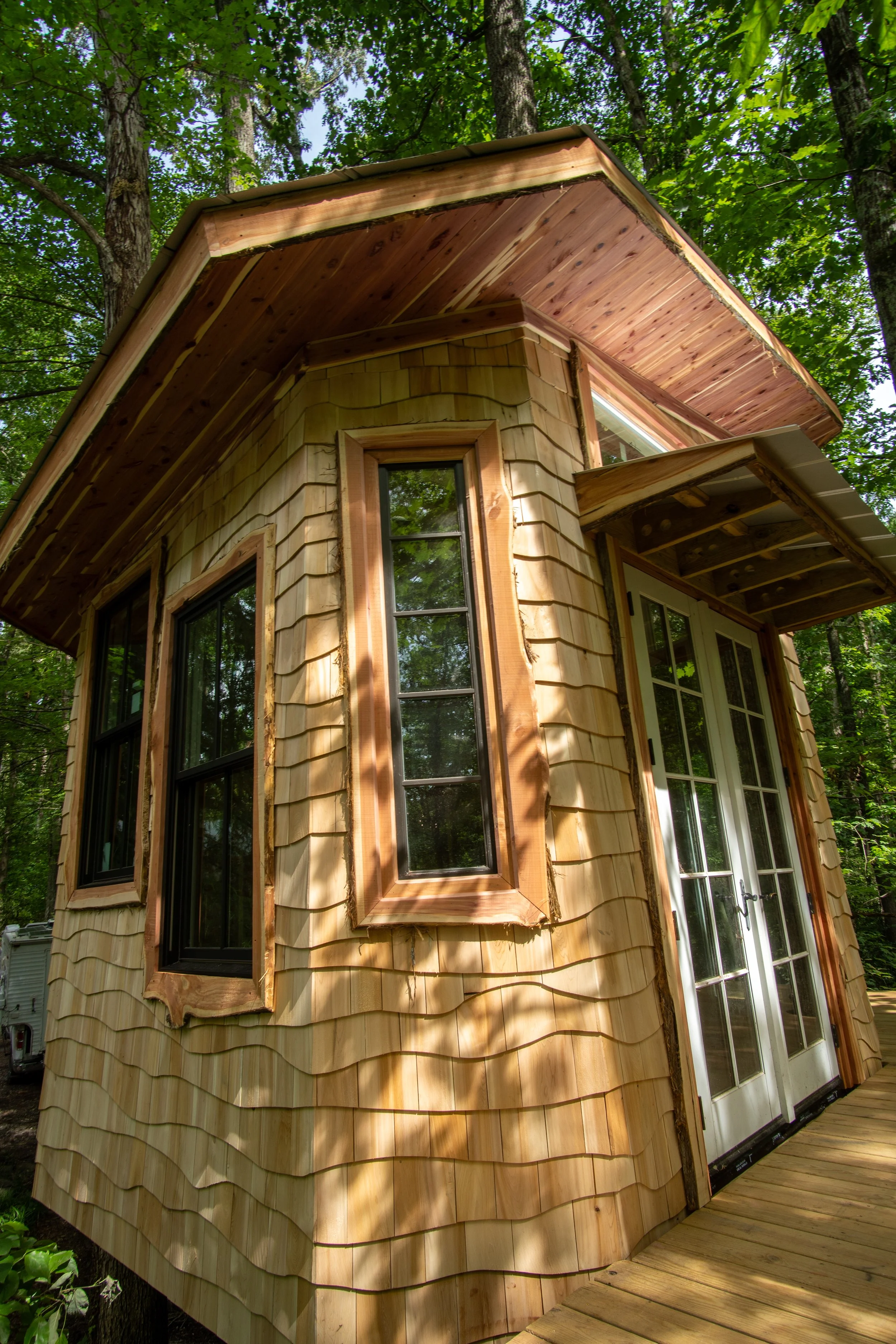 A small wooden house with shingle siding, black framed windows, a glass door, and a small overhang, situated among trees with lush green foliage.