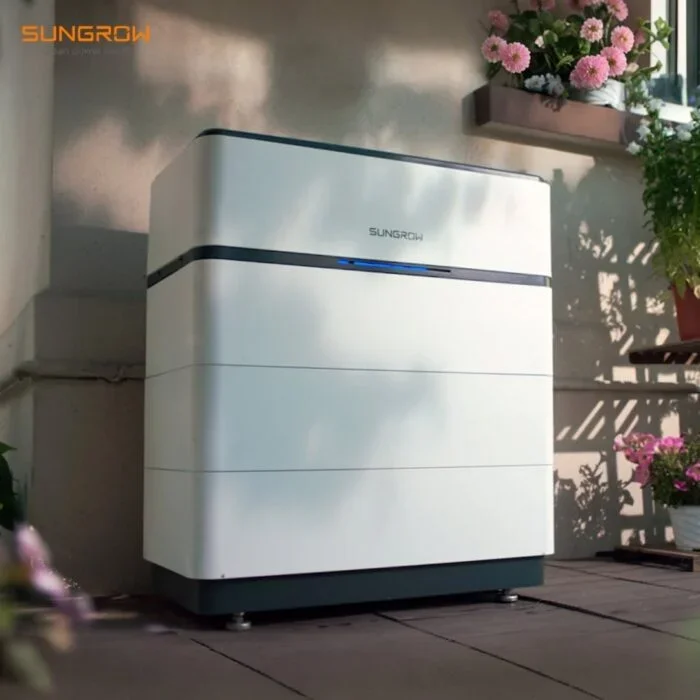 White portable air conditioning unit with blue indicator light, placed on a wooden floor near a wall with potted pink flowers and greenery.