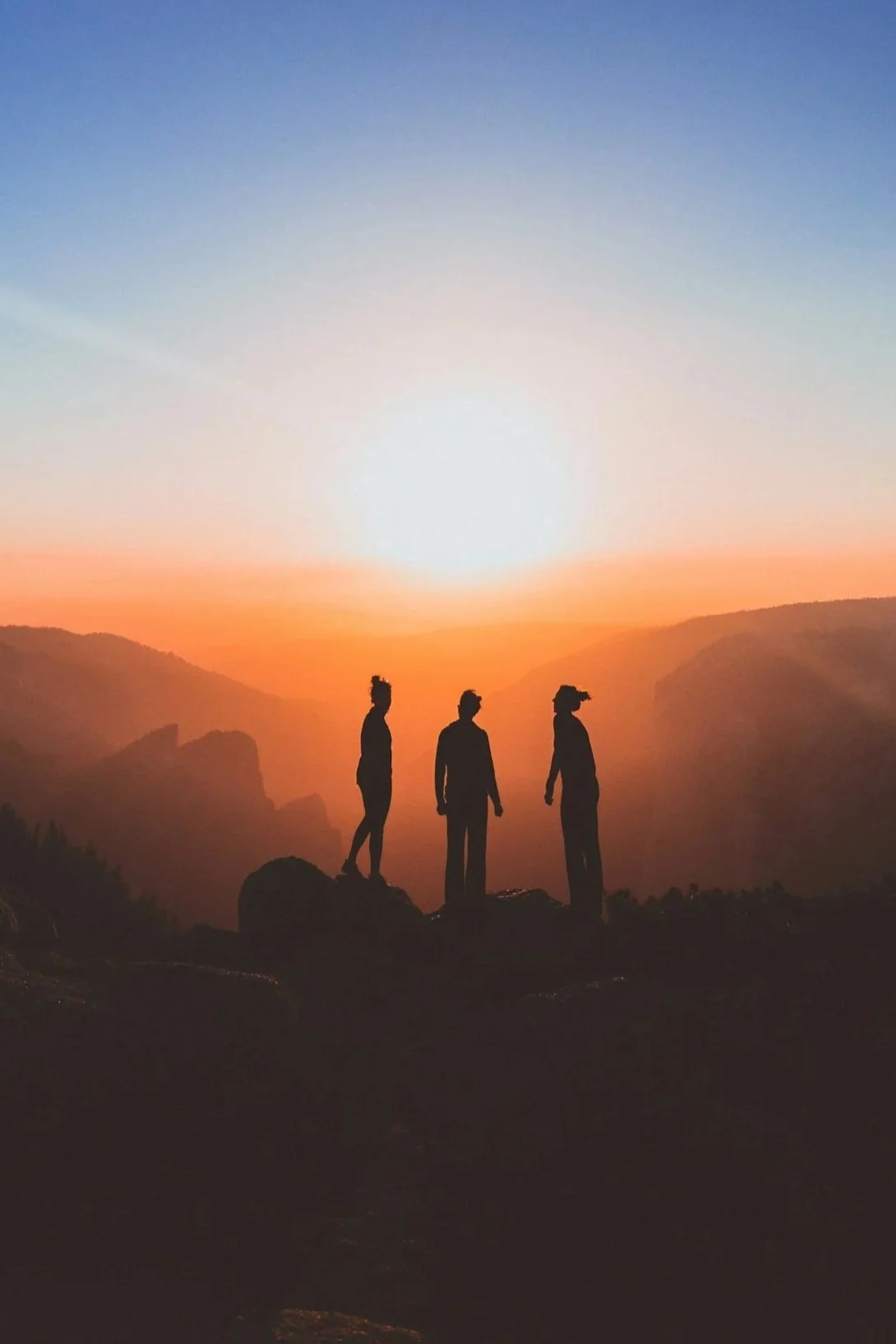Three women standing on rocks during sunset in a mountainous landscape.