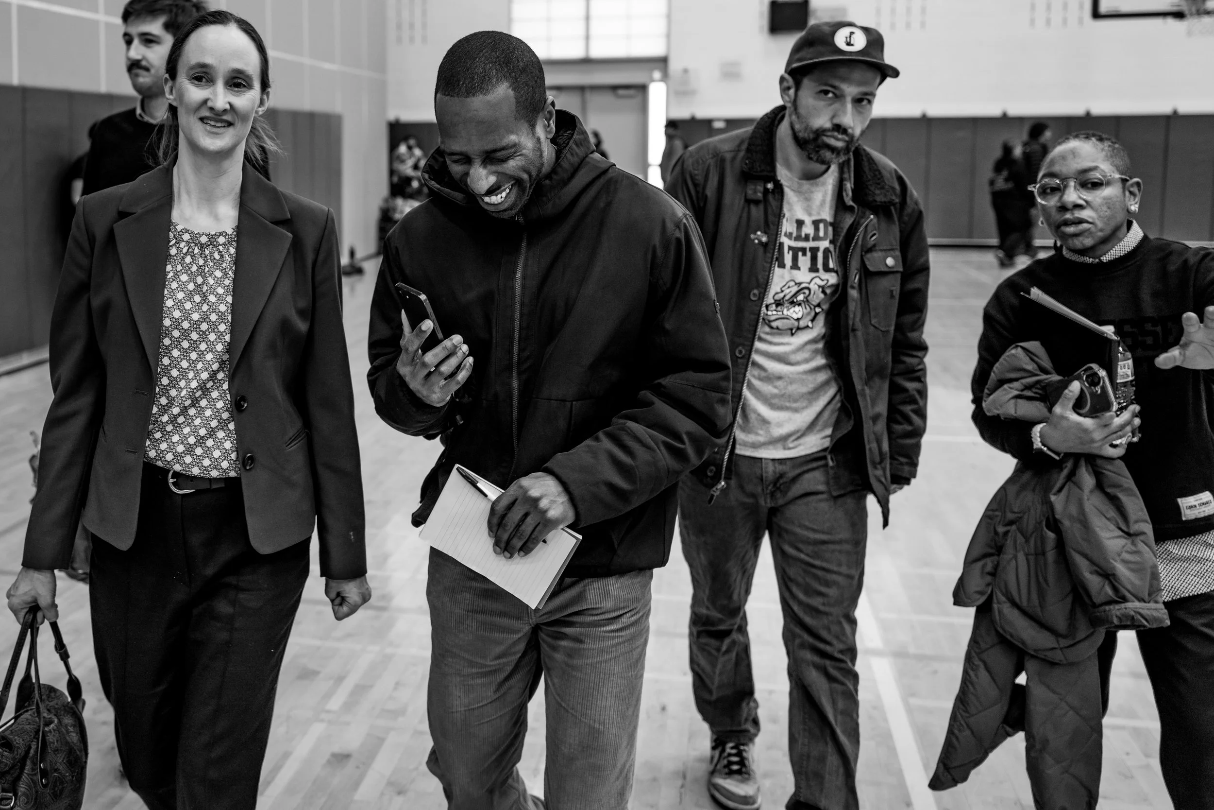Four people walking together indoors, two men and two women, with some holding notebooks and photo equipment, smiling and engaged in conversation.