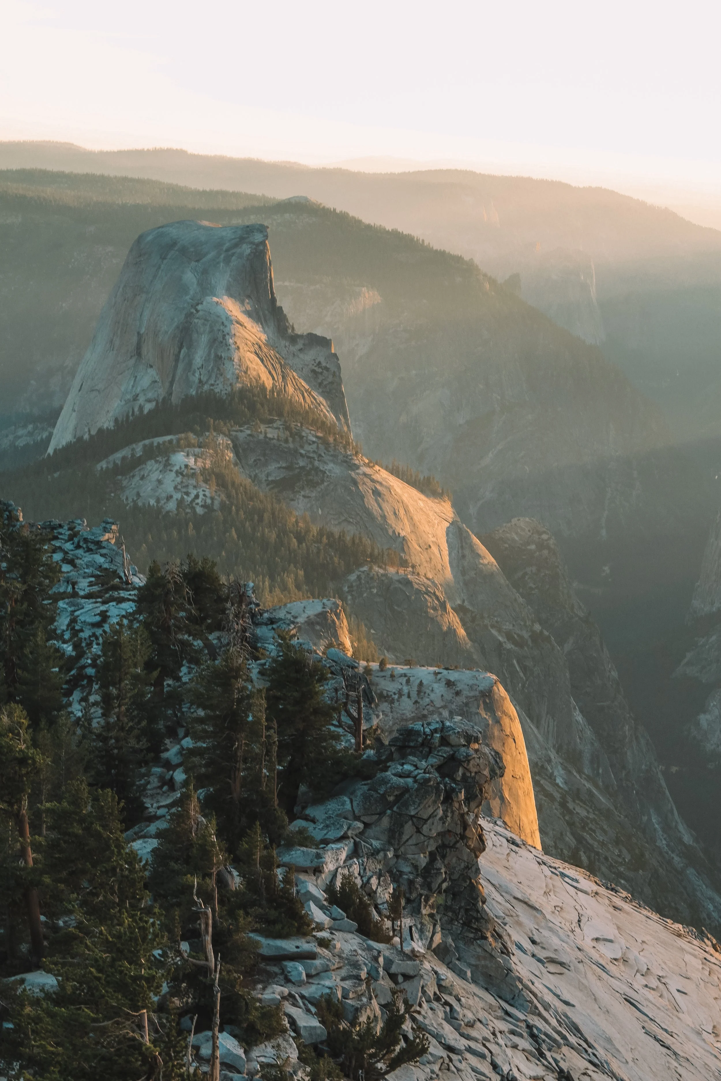 Sunlit view of Half Dome rock formation in Yosemite National Park with trees in foreground and mountain ranges in the background