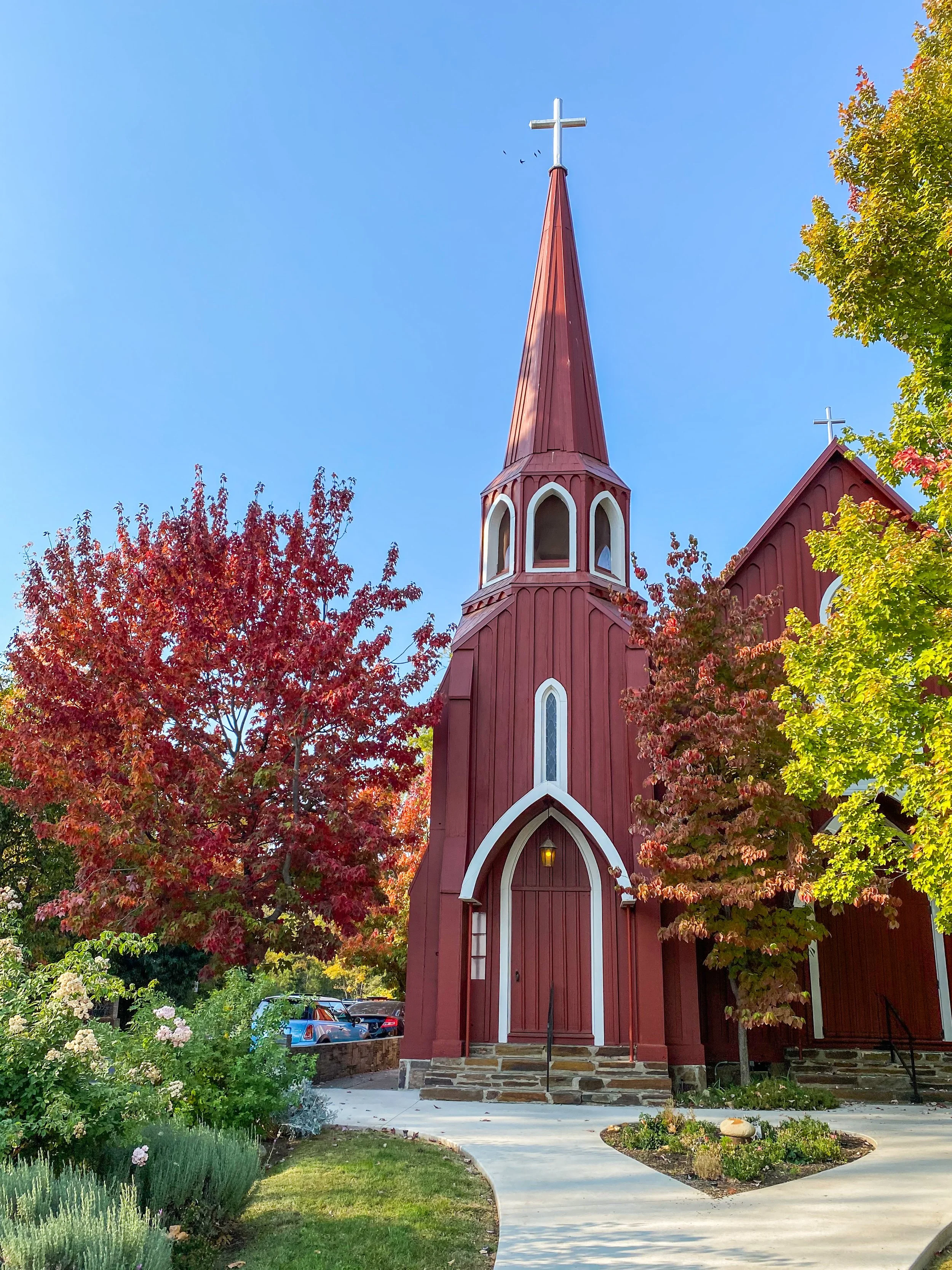 St. James Church in Sonora, California. A red wooden church with a tall steeple topped with a cross, surrounded by colorful autumn trees and a landscaped garden, under a clear blue sky.