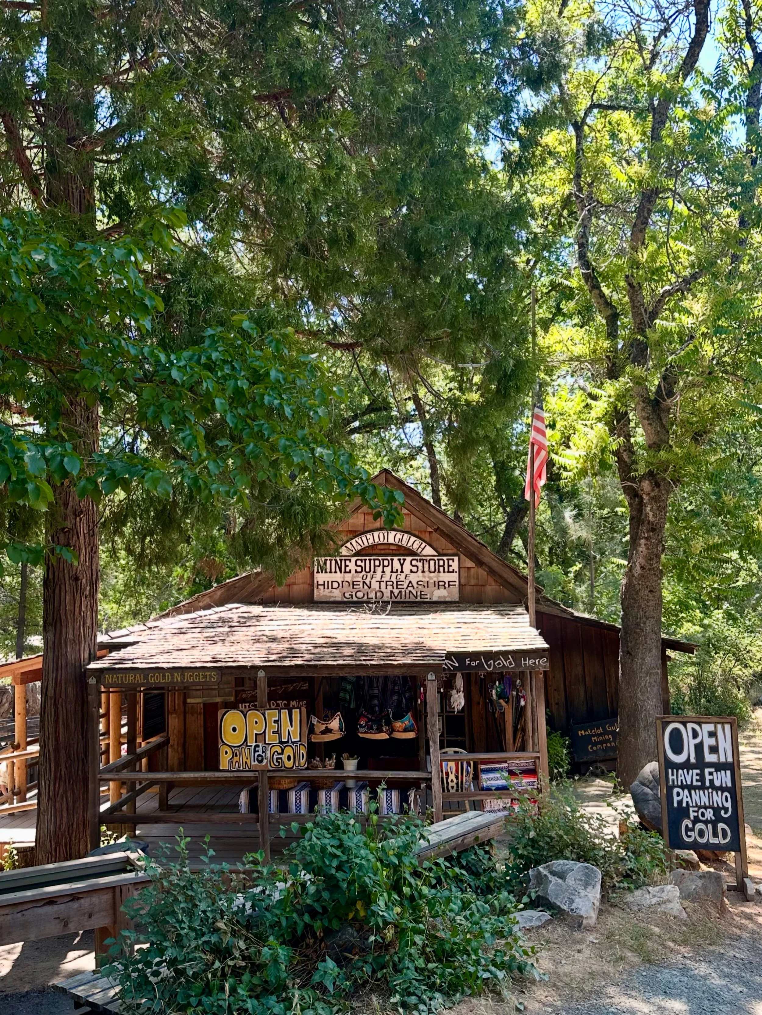 A small shop named Matelot Guch Mining Company in Columbia State Historic Park with signs indicating gold panning activities, surrounded by trees and foliage, with an American flag on a pole.