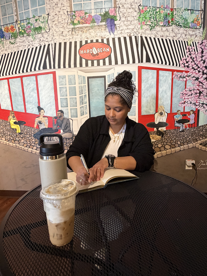 A woman sitting at a round table in a coffee shop, writing in a notebook with an iced coffee and a water bottle on the table, with a colorful mural of a cafe scene painted on the wall behind her.