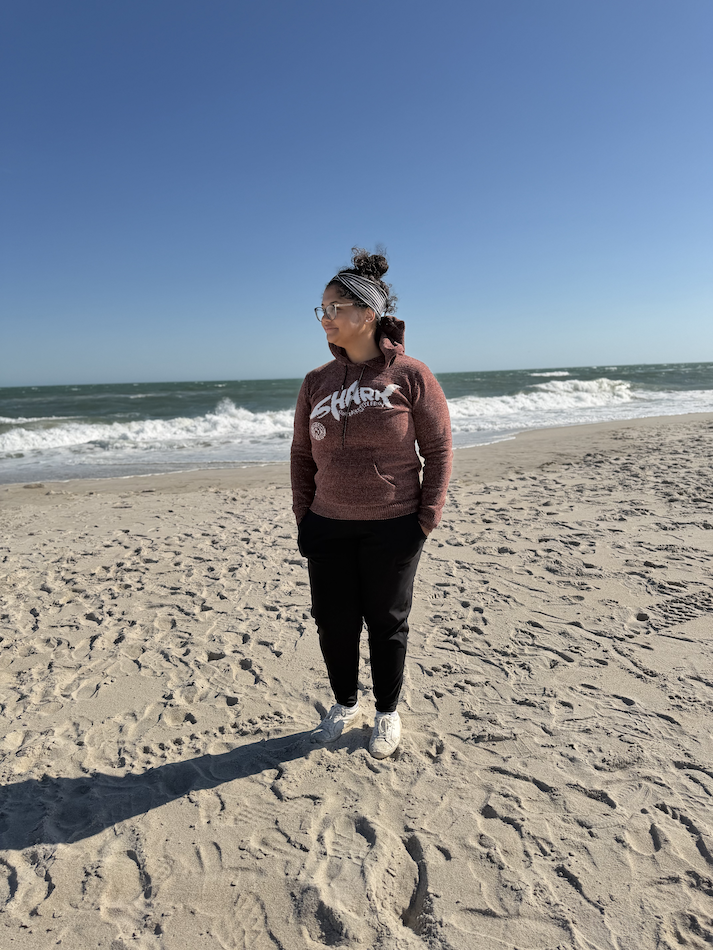 A woman standing on a sandy beach near the ocean, wearing a maroon hoodie, black pants, white sneakers, and glasses, with the sea and blue sky in the background.