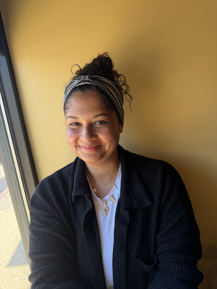 A woman with dark curly hair tied up, wearing a headband, black textured jacket, white shirt, and gold jewelry, sitting indoors next to a window with sunlight.
