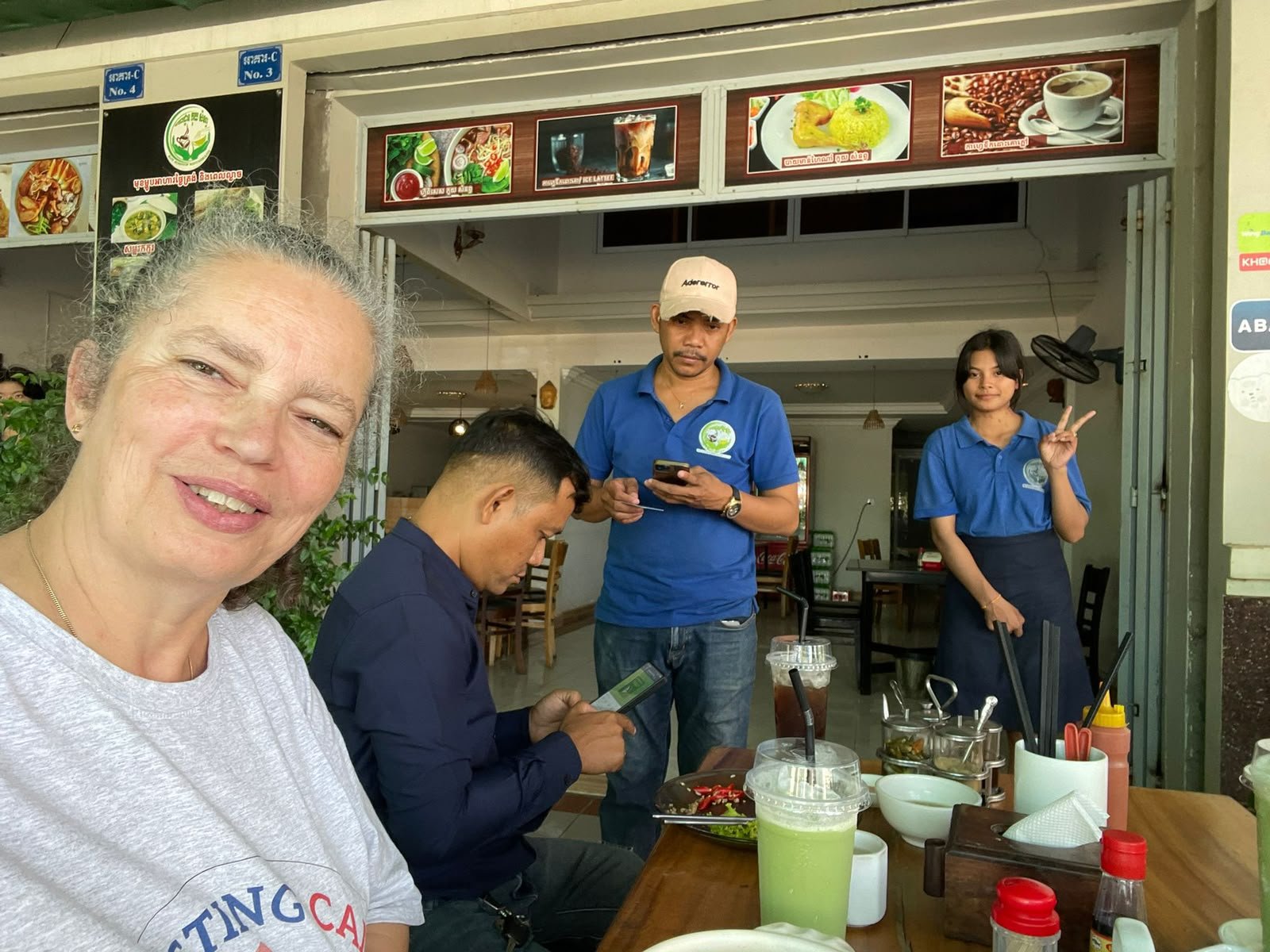 A woman smiling at the camera sits at a restaurant table with drinks and condiments. In the background, three restaurant staff members are standing, one of whom is making a peace sign, and another is looking at their phone. The restaurant has menu si
