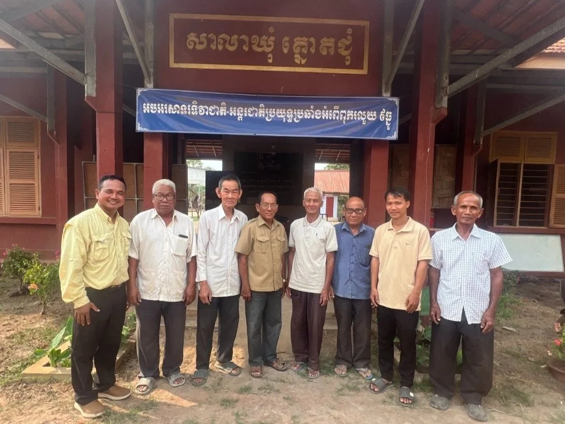 A group of eight men stands in a horizontal line for a photo in front of a traditional red wooden building with a tiled roof. A large blue banner with white Khmer script is centered above the group, and a motorcycle is parked on the right side of the