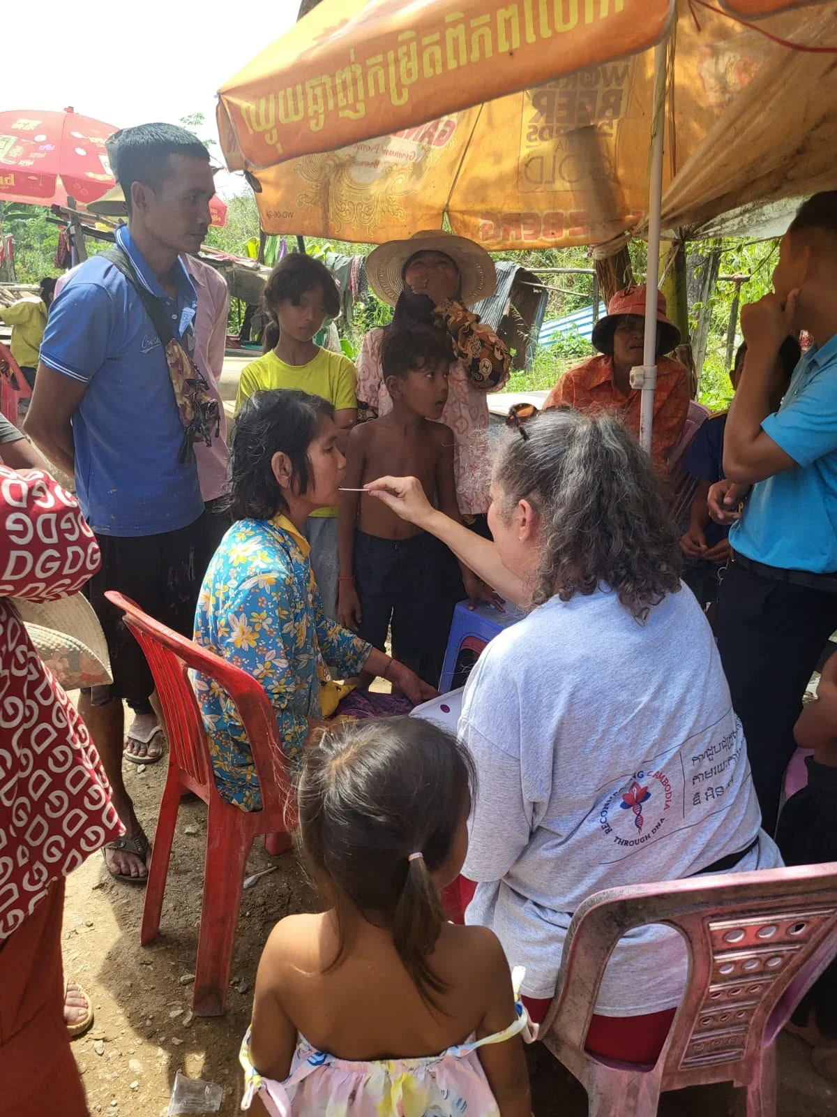 A woman receives a medical checkup from a healthcare worker, surrounded by children and adults under yellow and red umbrellas in a rural outdoor setting.