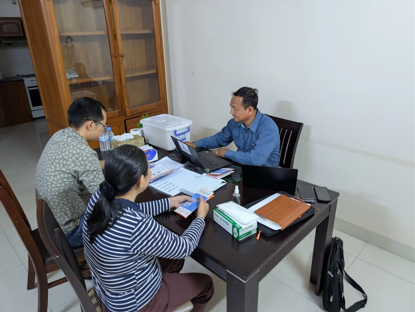 Three individuals are seated around a table engaged in a discussion or meeting. The table has various documents, electronic devices, and office supplies on it. The setting appears to be an office or meeting room with a plain white wall and a wooden c
