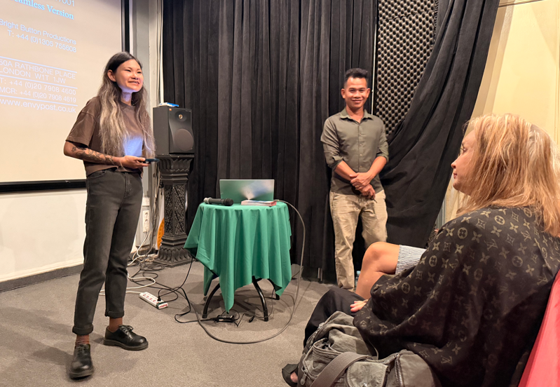 A woman stands at the front of a room speaking, with a presentation slide projected on the wall behind her. Two people stand nearby, listening, with a seated woman in the foreground observing the scene.