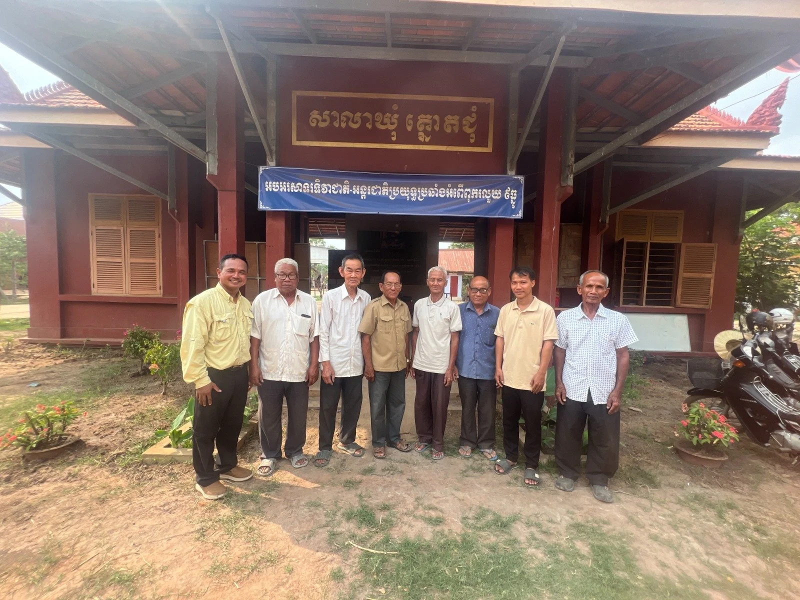 A group of nine men standing in front of a traditional building with red walls and a tiled roof. There are potted plants on either side of the group and motorcycles to the right. The men are dressed in casual and semi-formal attire, smiling at the ca