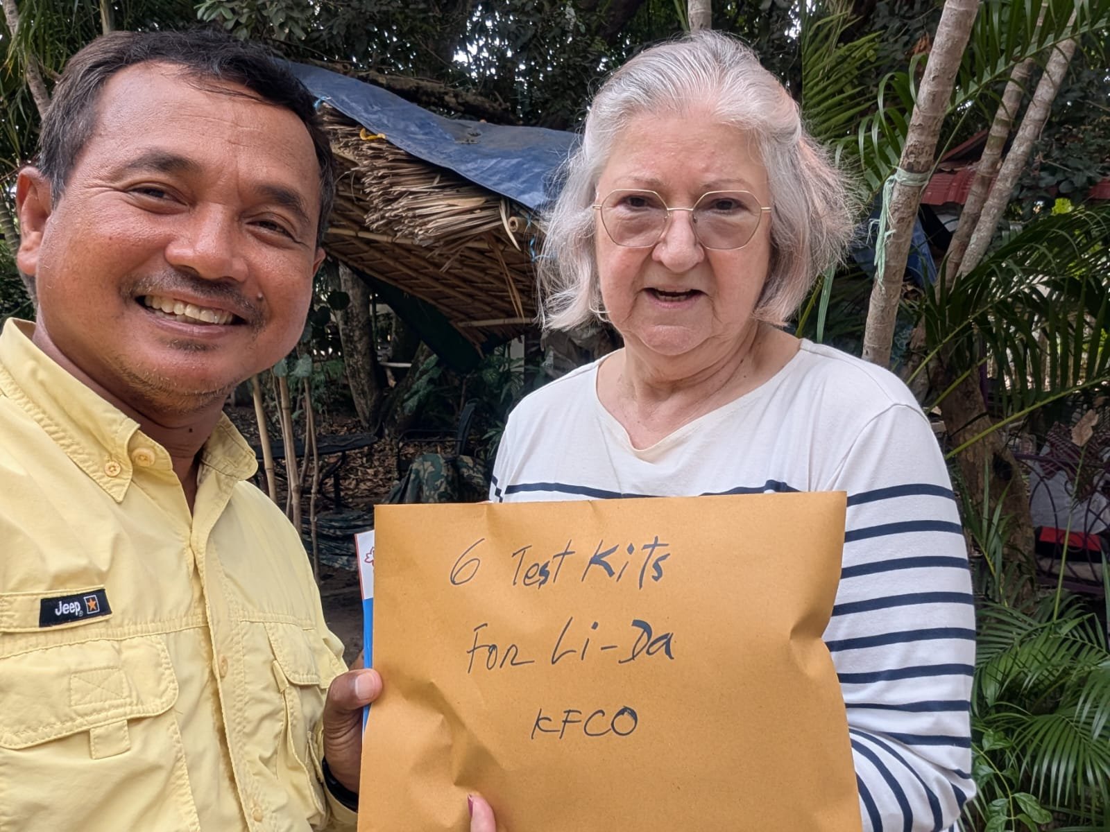 A man and an elderly woman outdoors, smiling at the camera. The woman holds a sign that reads '6 Test Kits for Li-Da KECO'. There are trees and a rustic wooden structure in the background.