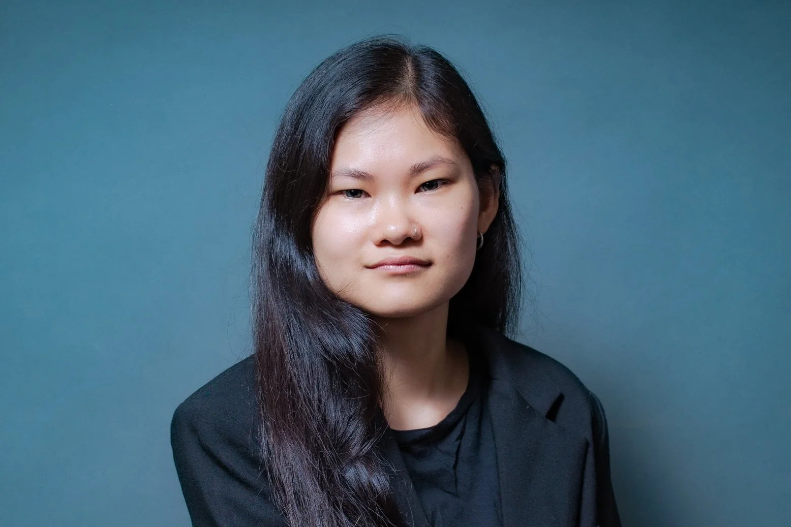 Headshot of a woman with long dark hair, wearing a black blazer, against a plain blue background.