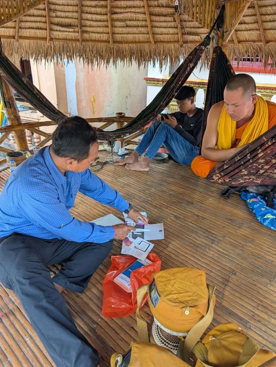 Man in a blue shirt showing documents to a monk in orange robes, while a young person sitting in a hammock looks at a phone, under a thatched roof structure.