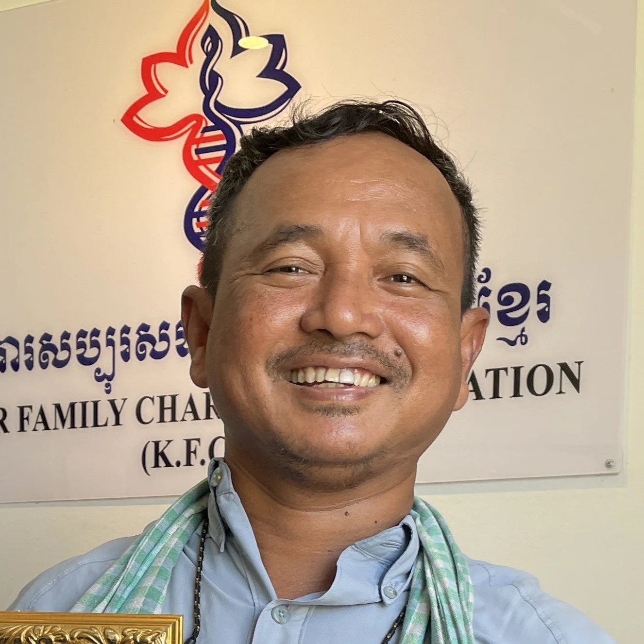 Smiling man in front of a sign for a family charity foundation with a logo of a flower and DNA strand, wearing a light blue shirt and a striped scarf.